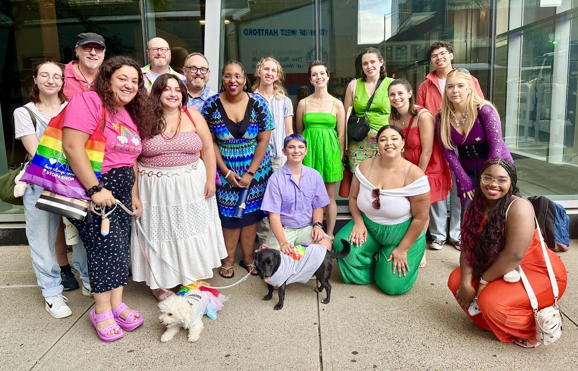 Group of diverse people gathered outside a building, some wearing colorful attire, with two dogs in front.