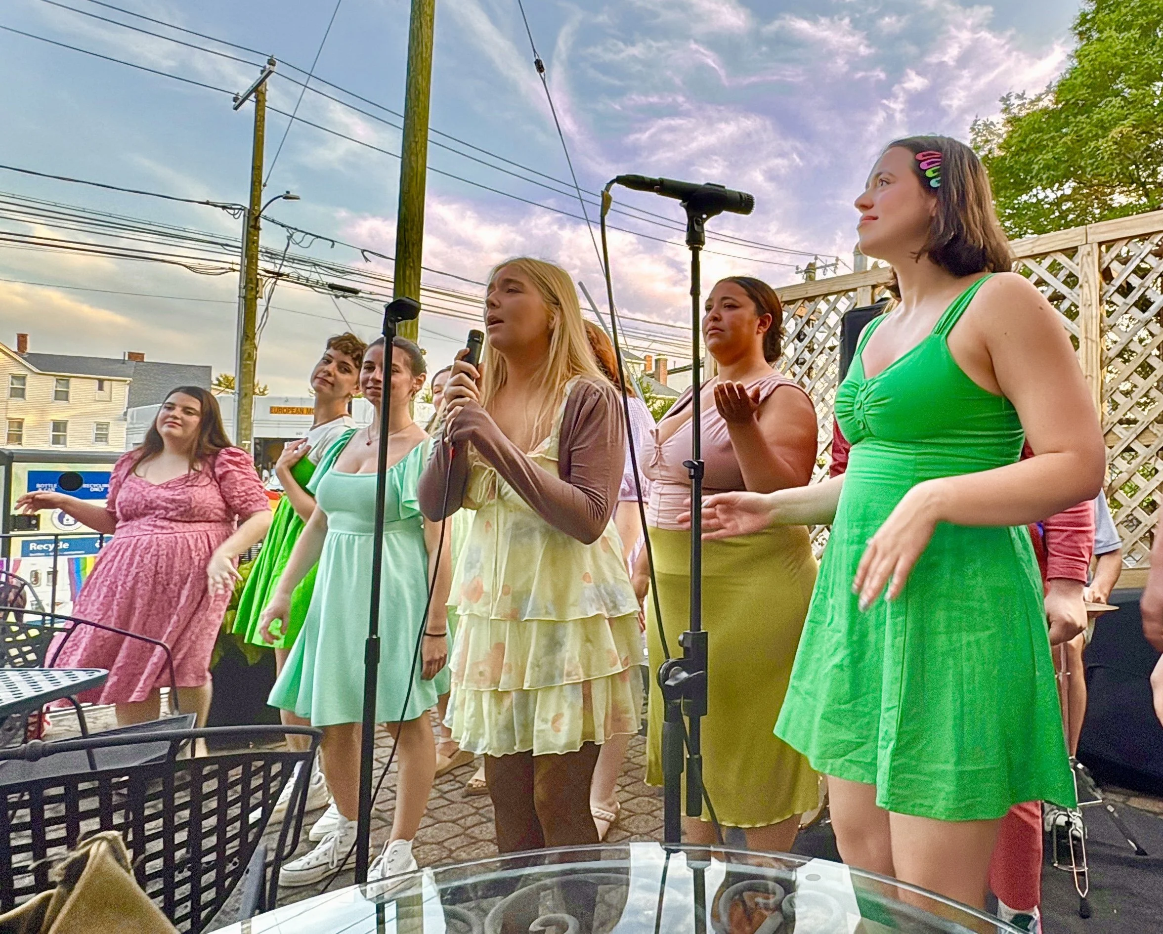 A group of women singing outdoors at sunset, some with microphones, wearing colorful dresses, with a wooden fence, power lines, and houses in the background.