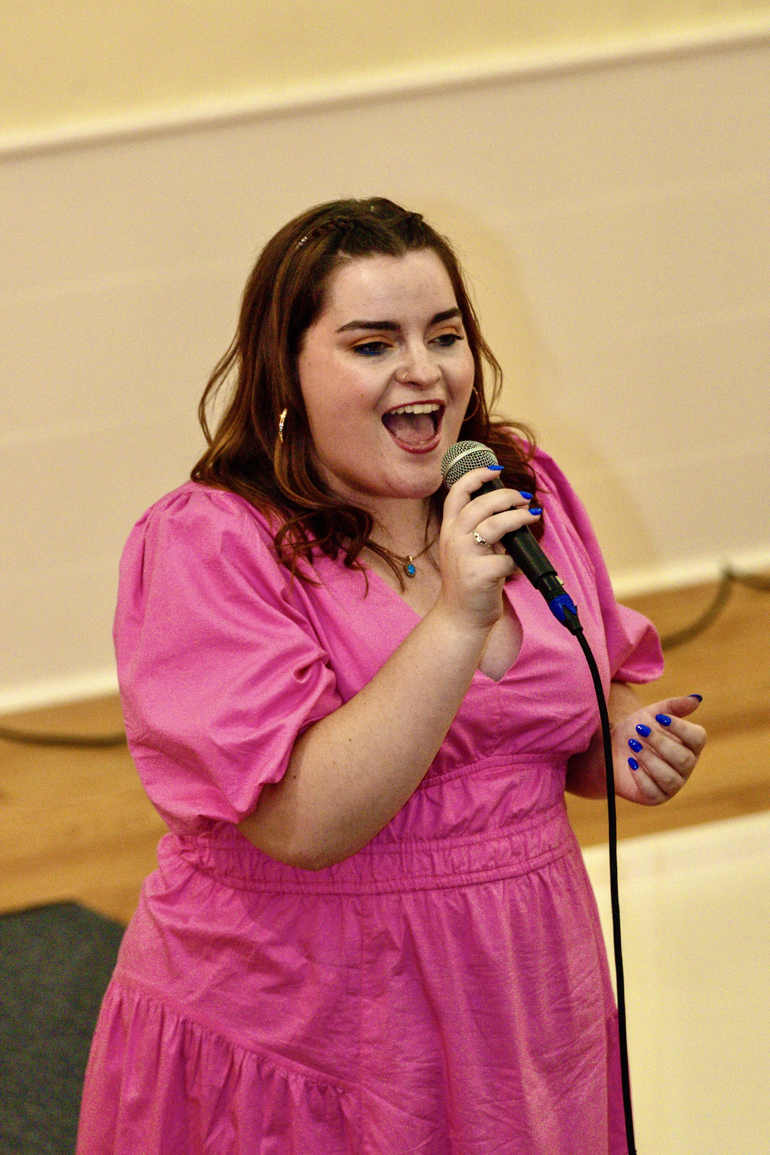 A woman with brown hair, wearing a pink dress and jewelry, singing into a microphone.