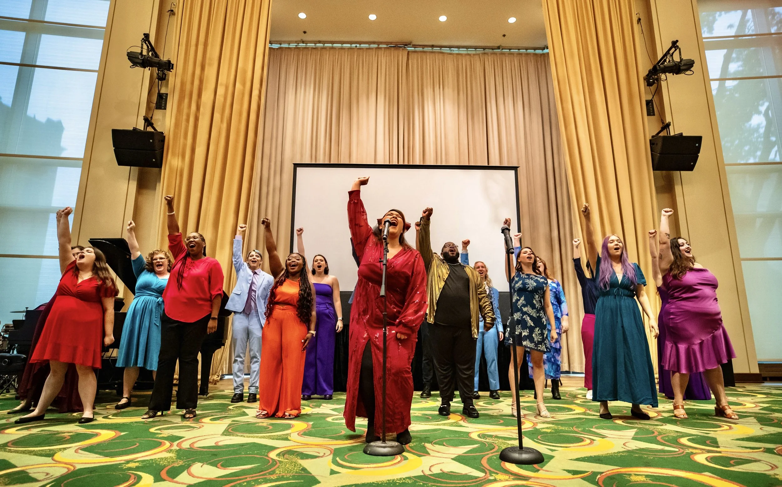 A diverse group of performers on stage raising their fists, with the central singer in red, during a concert or performance in a grand hall with gold curtains and a large screen behind them.