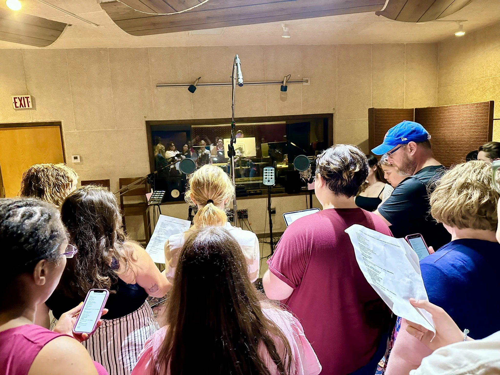 People in a recording studio, some holding sheets of paper and smartphones, with microphones and sound equipment in the background.