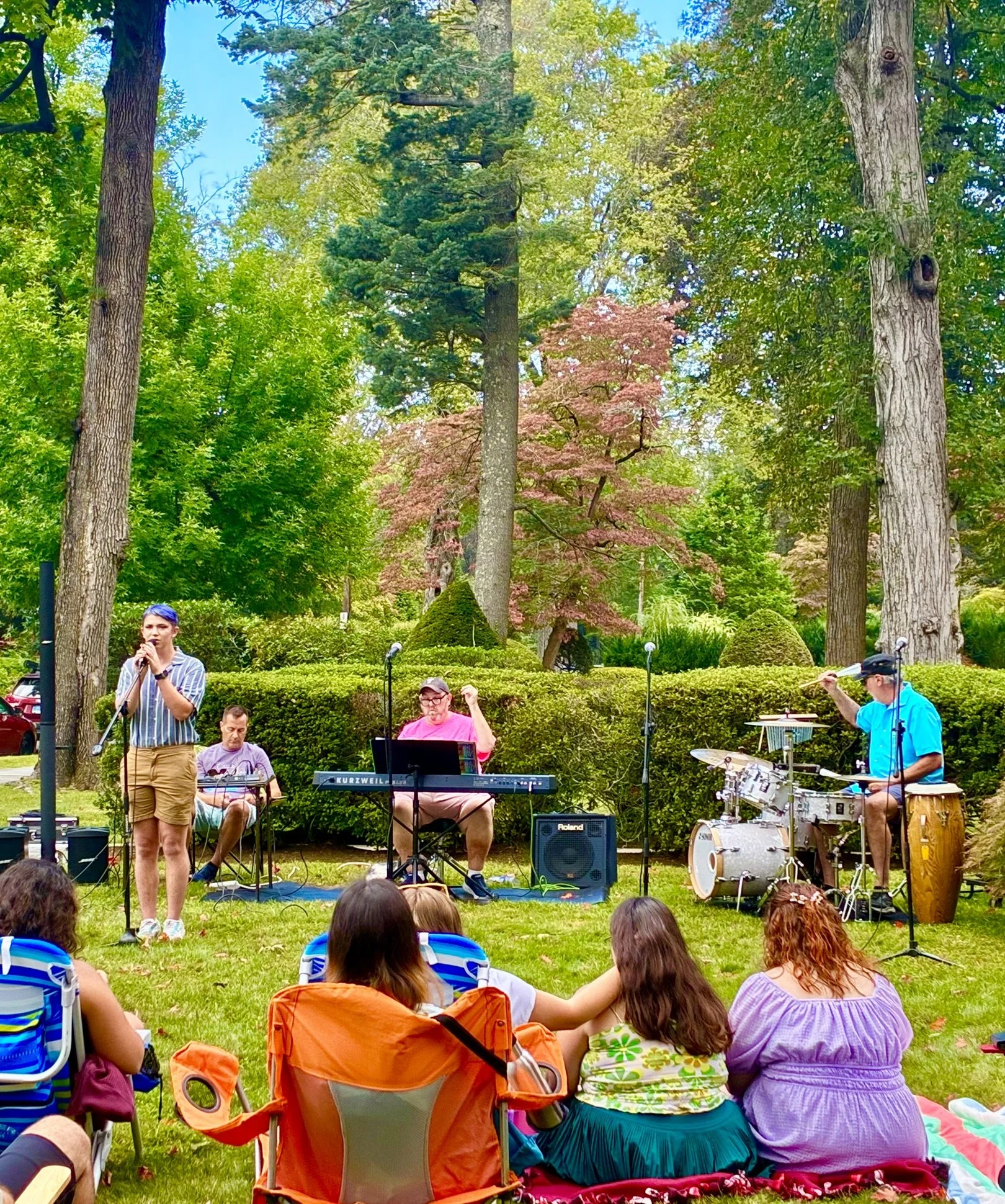 A live outdoor music performance in a park with trees and greenery, featuring a female singer, a keyboardist, a bassist, and a drummer, with an audience sitting on blankets and chairs.