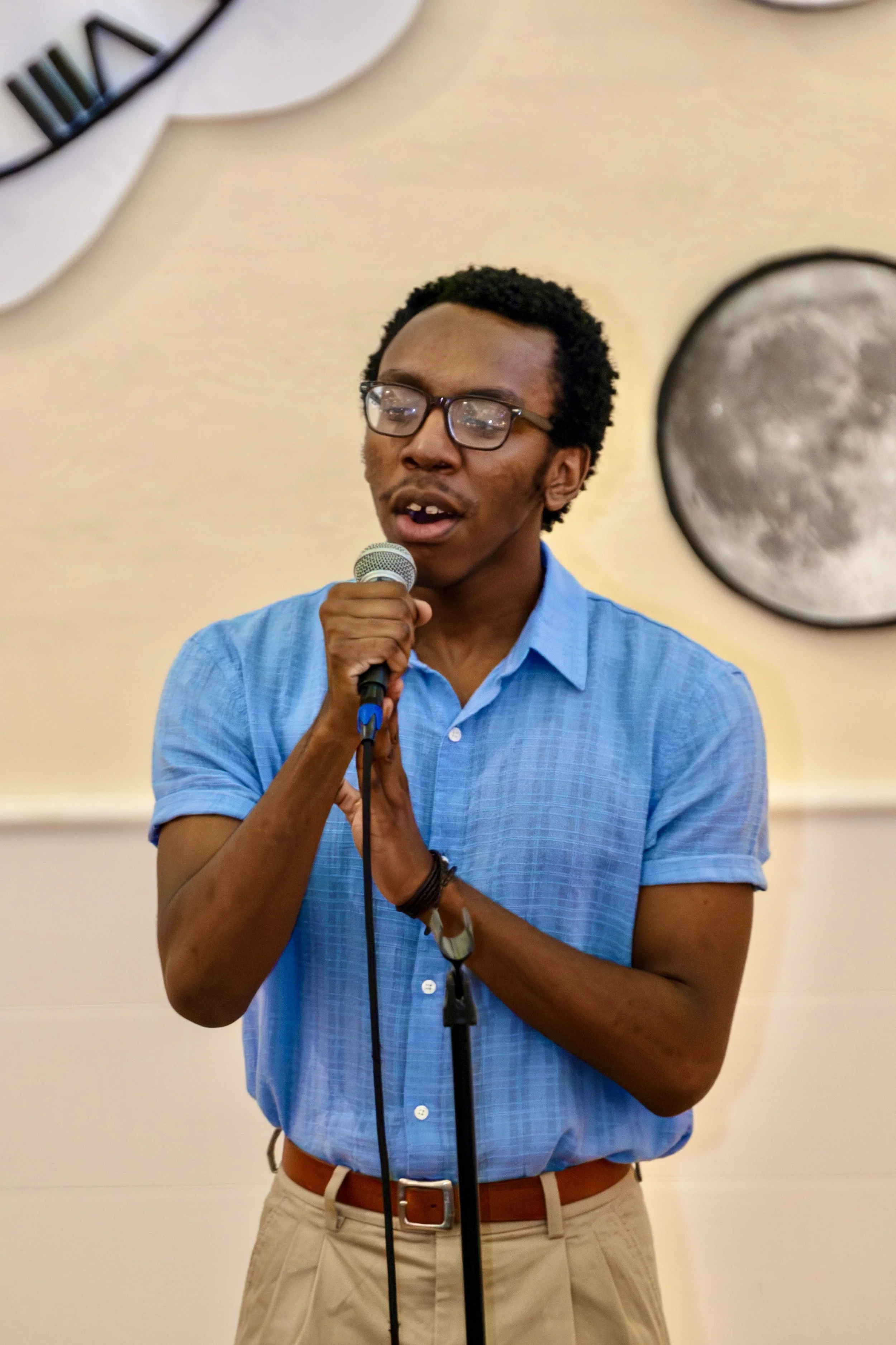 A man wearing glasses, a blue short-sleeved shirt, and khaki pants speaking into a microphone.
