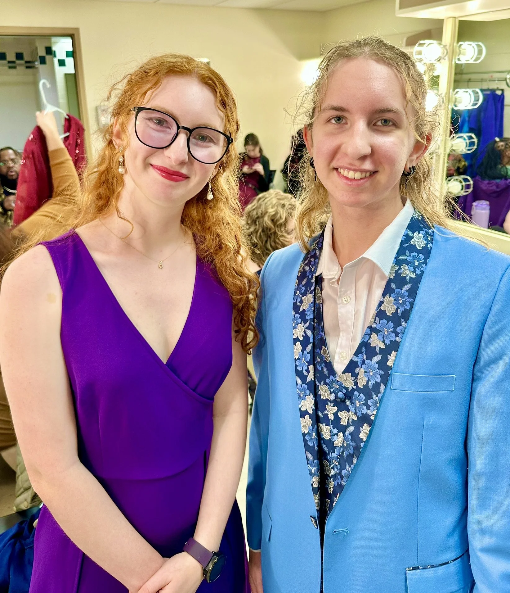 Two women smiling at a social event, with one wearing a purple dress and glasses, and the other in a blue blazer with a floral scarf. In the background, people are in a dressing room or backstage area.