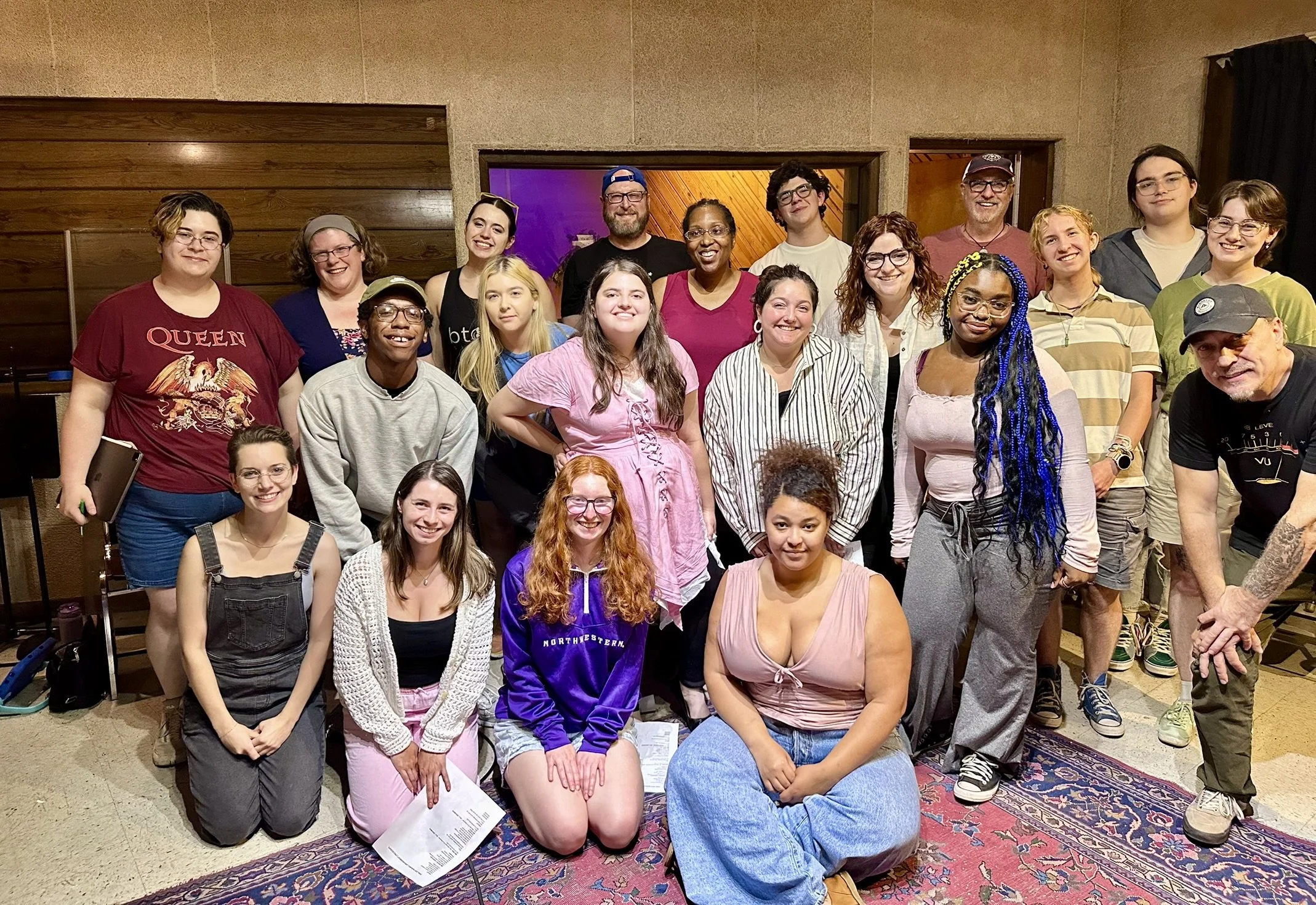 Group of 21 diverse people posing together indoors, some kneeling in front, others standing behind, in a room with wooden and beige walls, with a purple-lit area in the background.
