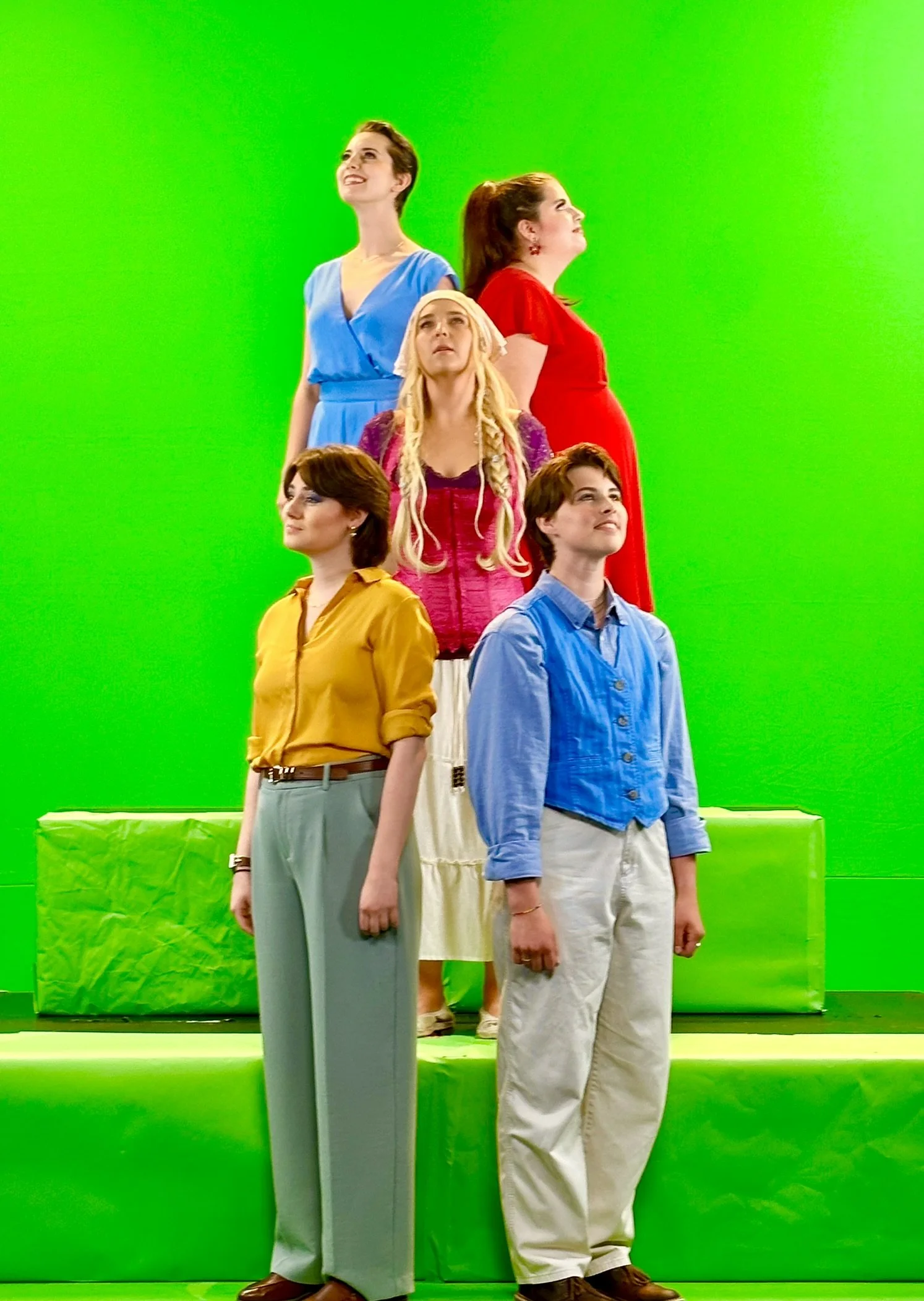 Six women standing on green screens in multiple rows, dressed in colorful clothing, looking upward.