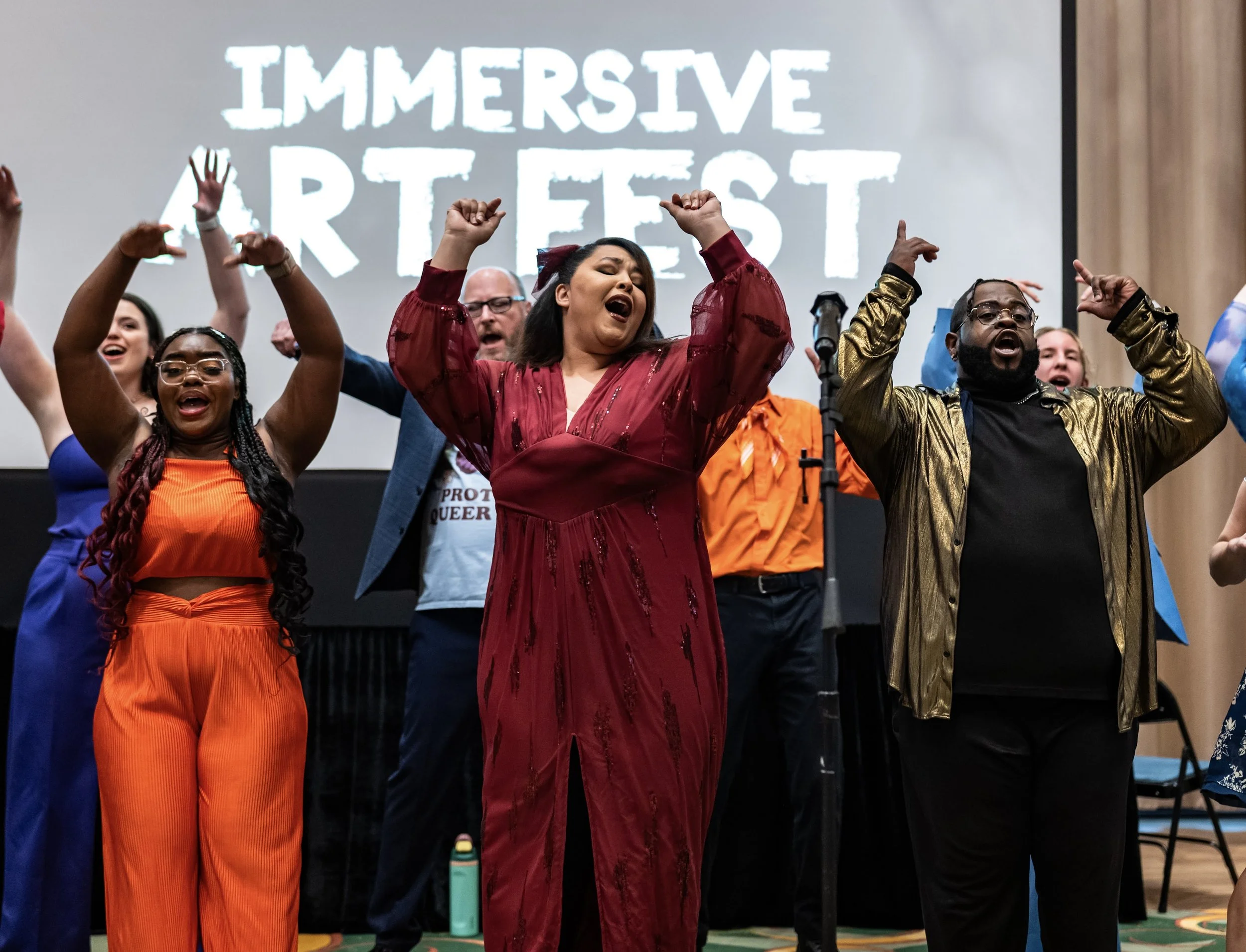 Group of diverse people singing and dancing on stage during an event, with a large screen in the background displaying "IMMERSIVE ART FEST."