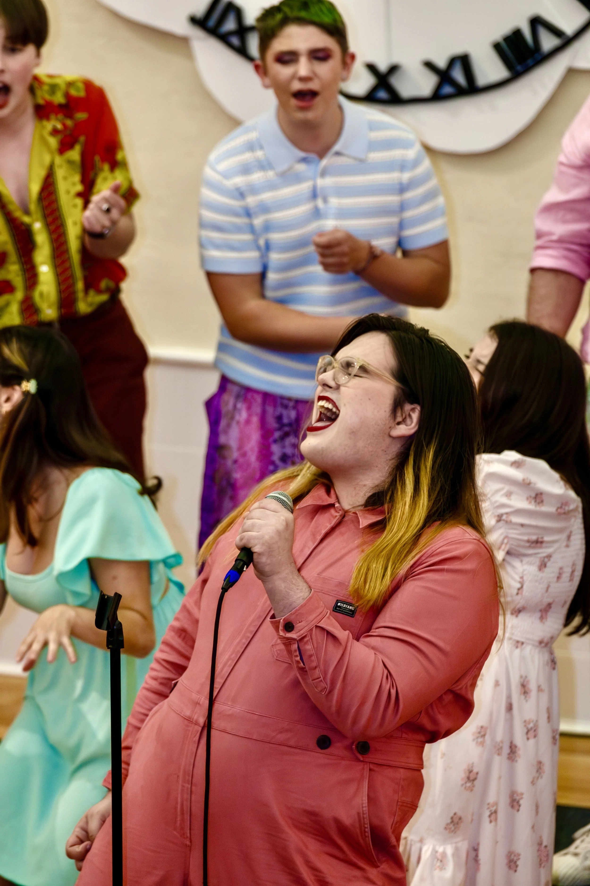 A woman with glasses and blonde hair with black roots, wearing a pink jacket, is singing joyfully into a microphone at a social gathering. Other people are gathered around her, with some enjoying the moment, and a large clock is visible on the wall b