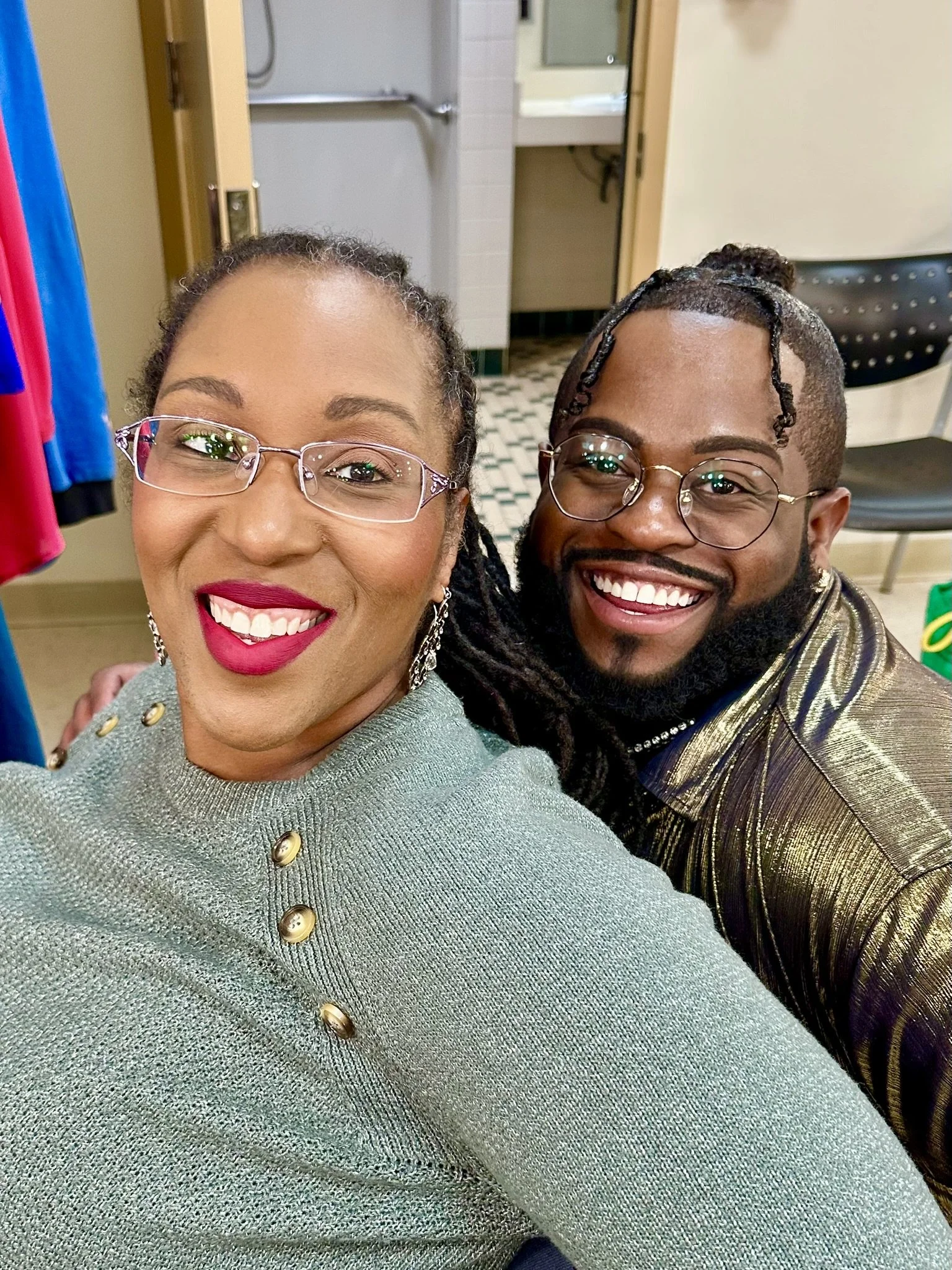 A smiling woman with glasses and bright lipstick takes a selfie with a smiling man with glasses and dreadlocks. They are indoors, possibly in a community center or meeting space, with chairs and a bathroom in the background.