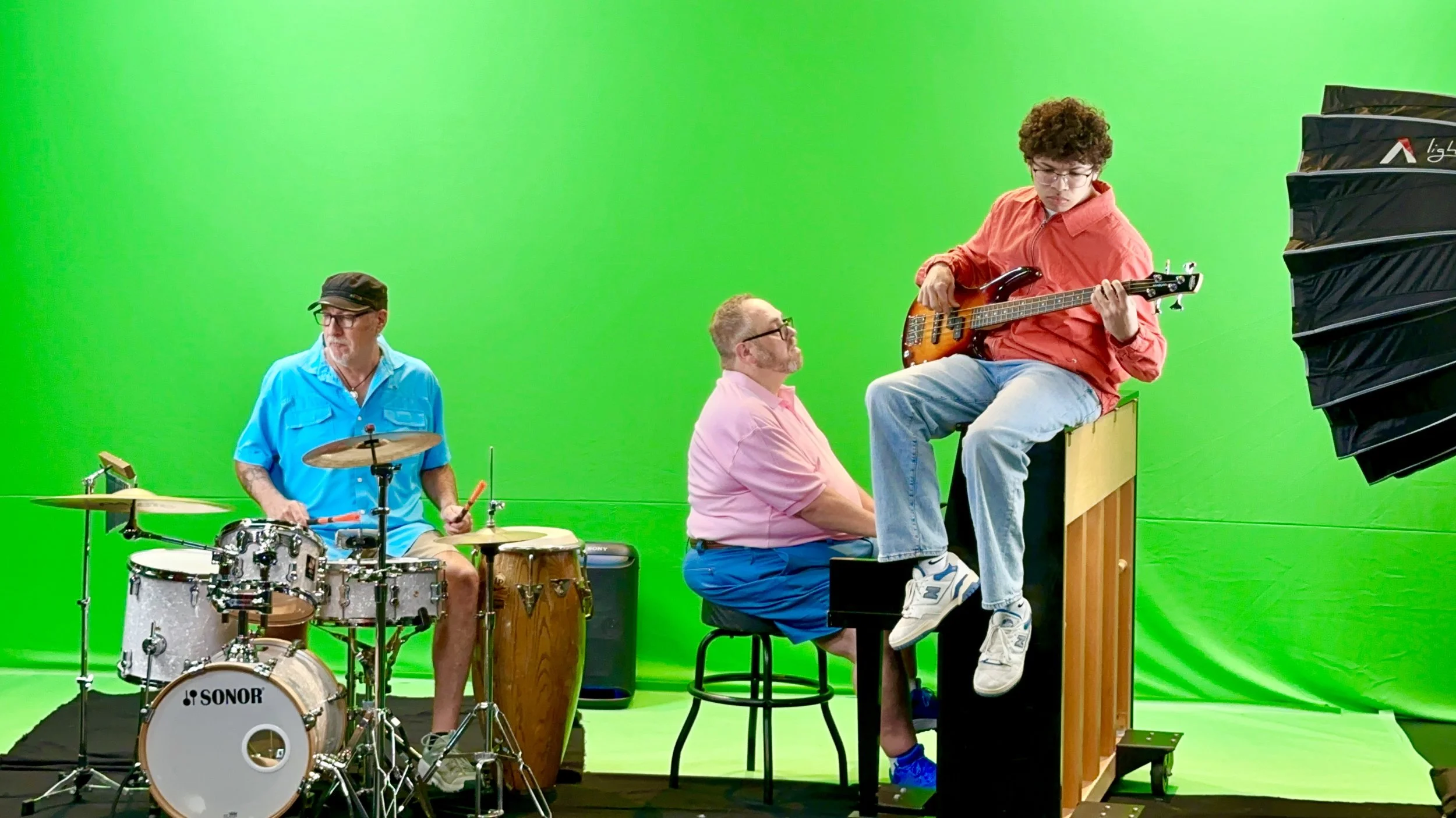 Three men playing musical instruments in front of a green screen: one on drums, one on a keyboard, and one on a bass guitar.