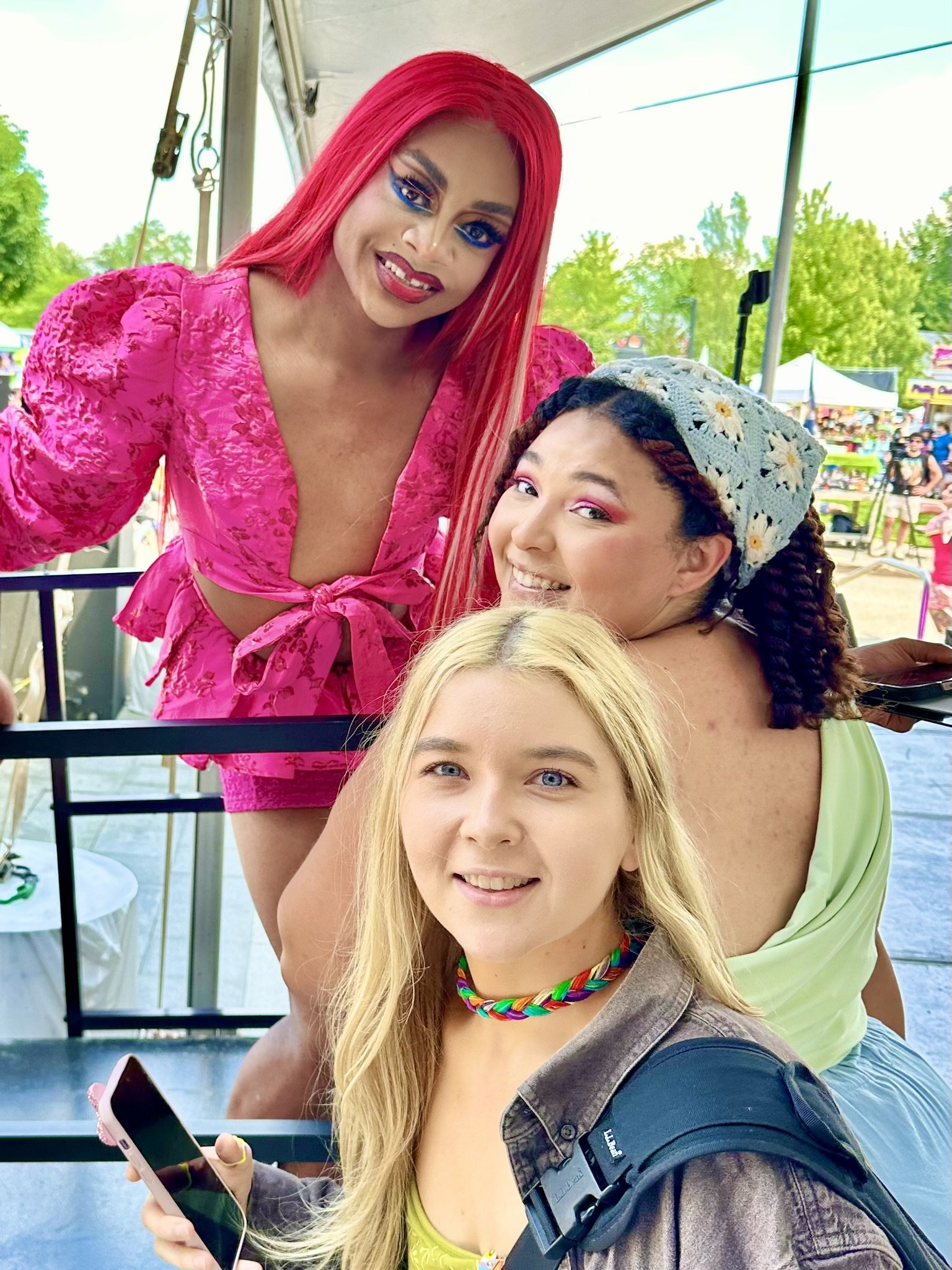 Three women at an outdoor event; one with bright pink hair and a pink dress, one with curly hair and a floral headband, and one with long blonde hair holding a phone, smiling.