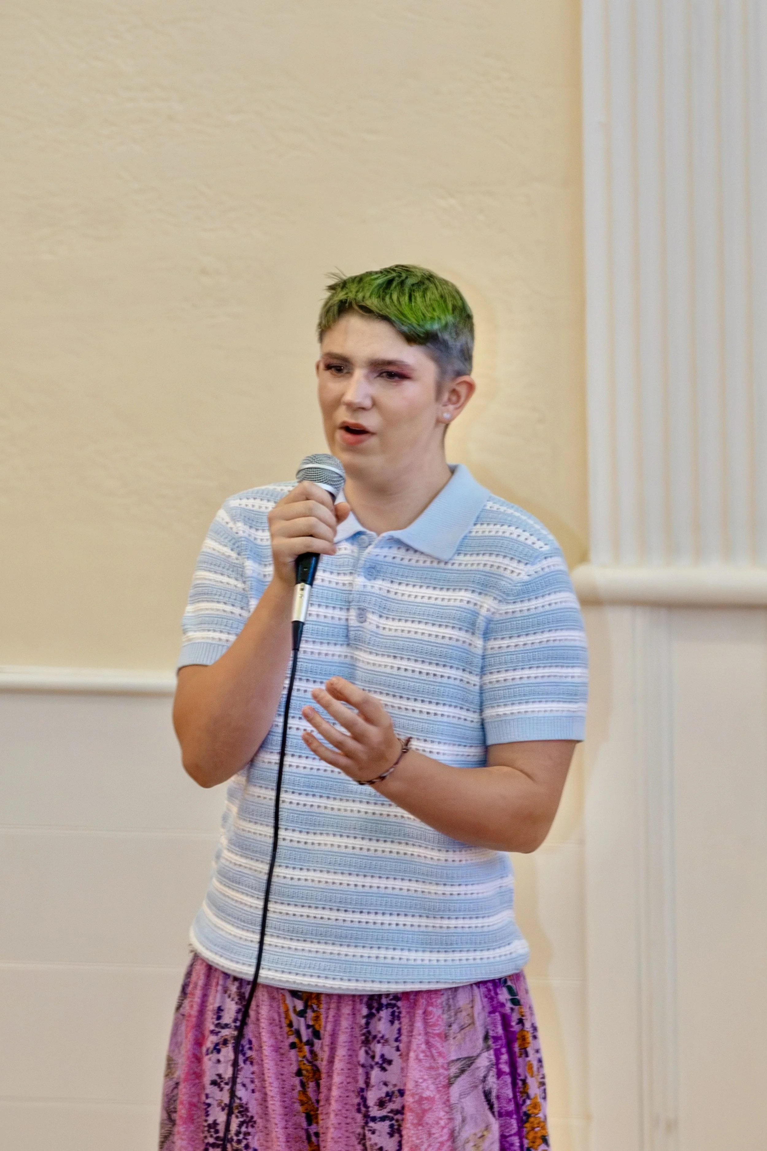 A person with short hair dyed green on top, speaking into a microphone.