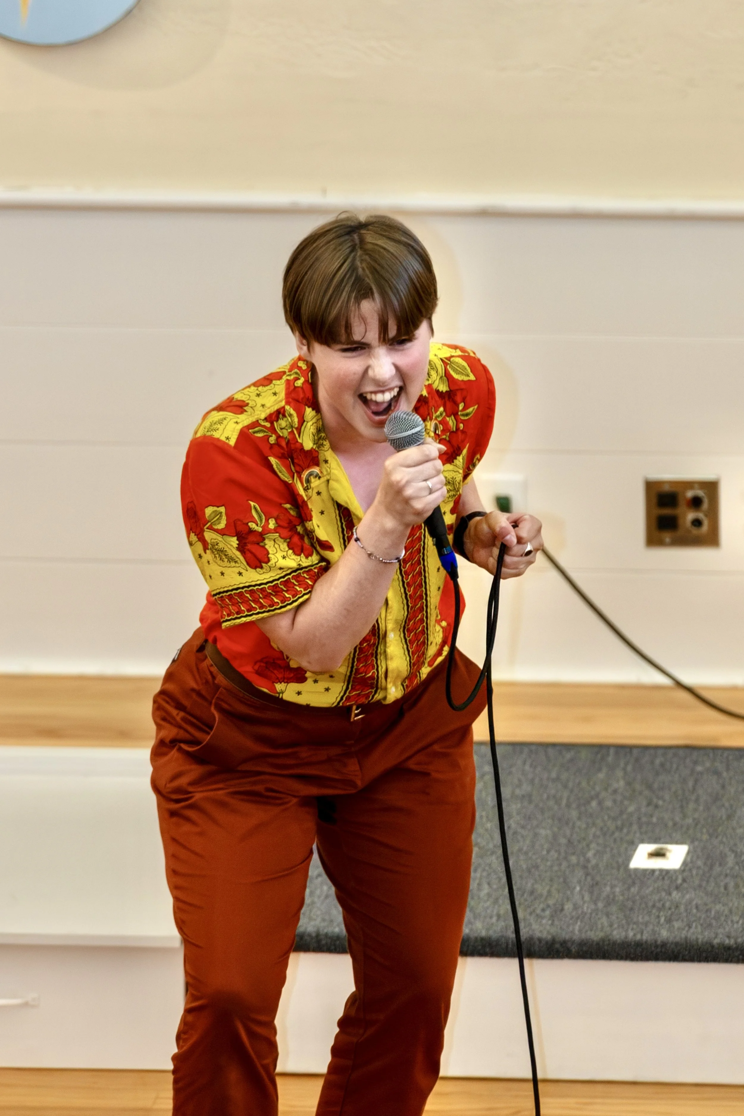 A person singing passionately into a microphone on stage, wearing a vibrant red and yellow patterned shirt and orange pants.