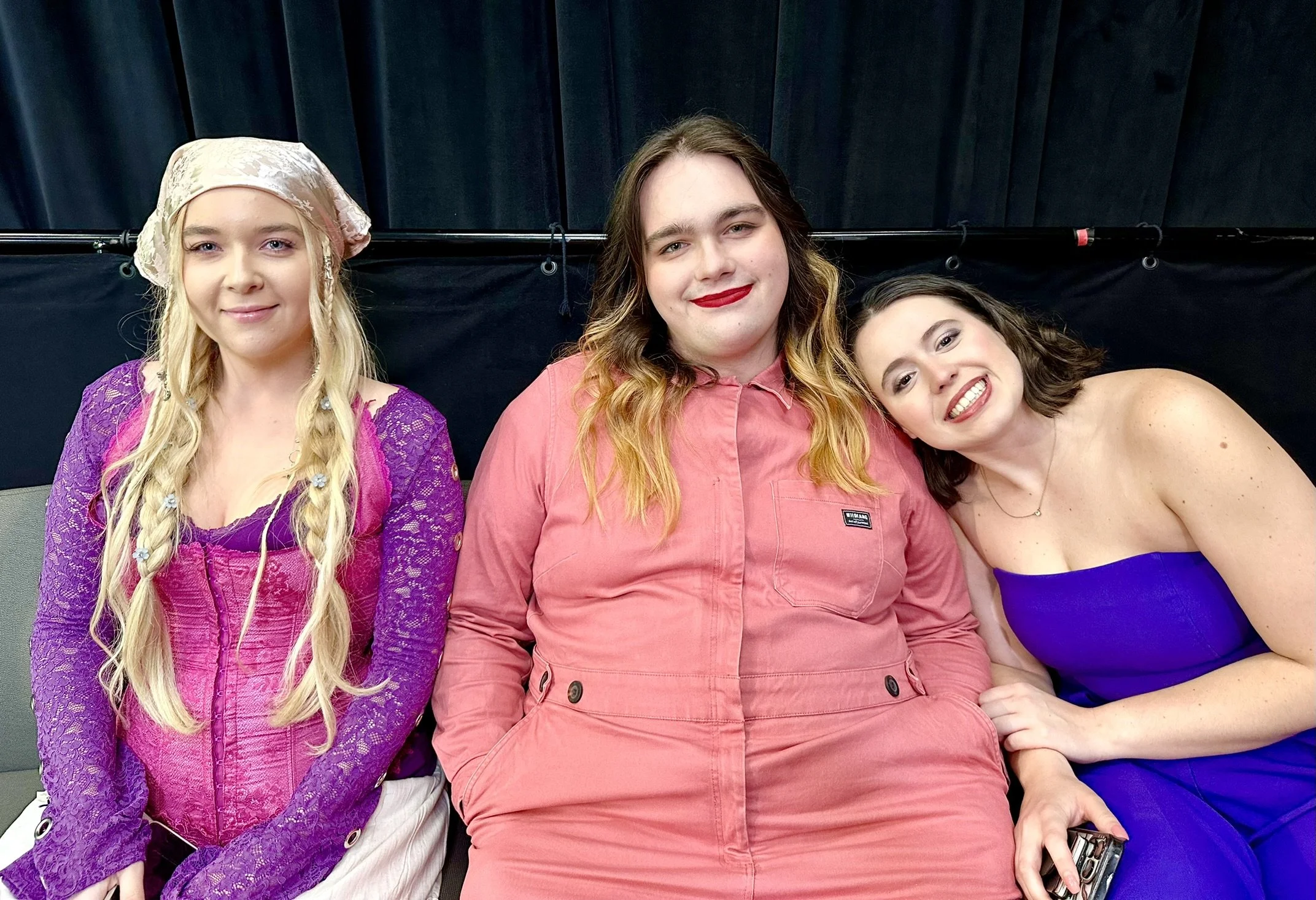 Three women sitting together, smiling, against a black curtain backdrop. The woman on the left has long blonde hair, a pink and purple outfit with a lace top, and a pink head covering. The woman in the middle has long wavy brown hair, wears a pink ju