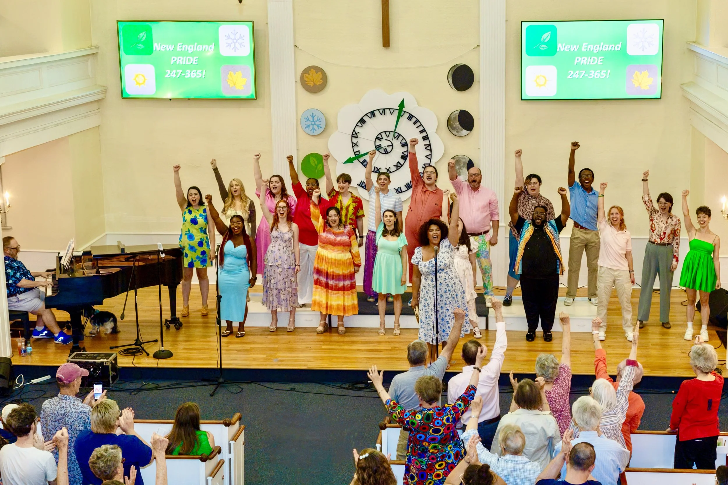 A diverse group of people standing on stage with their fists raised, during a singing or speaking event at a church or community hall, with an audience in front of them.