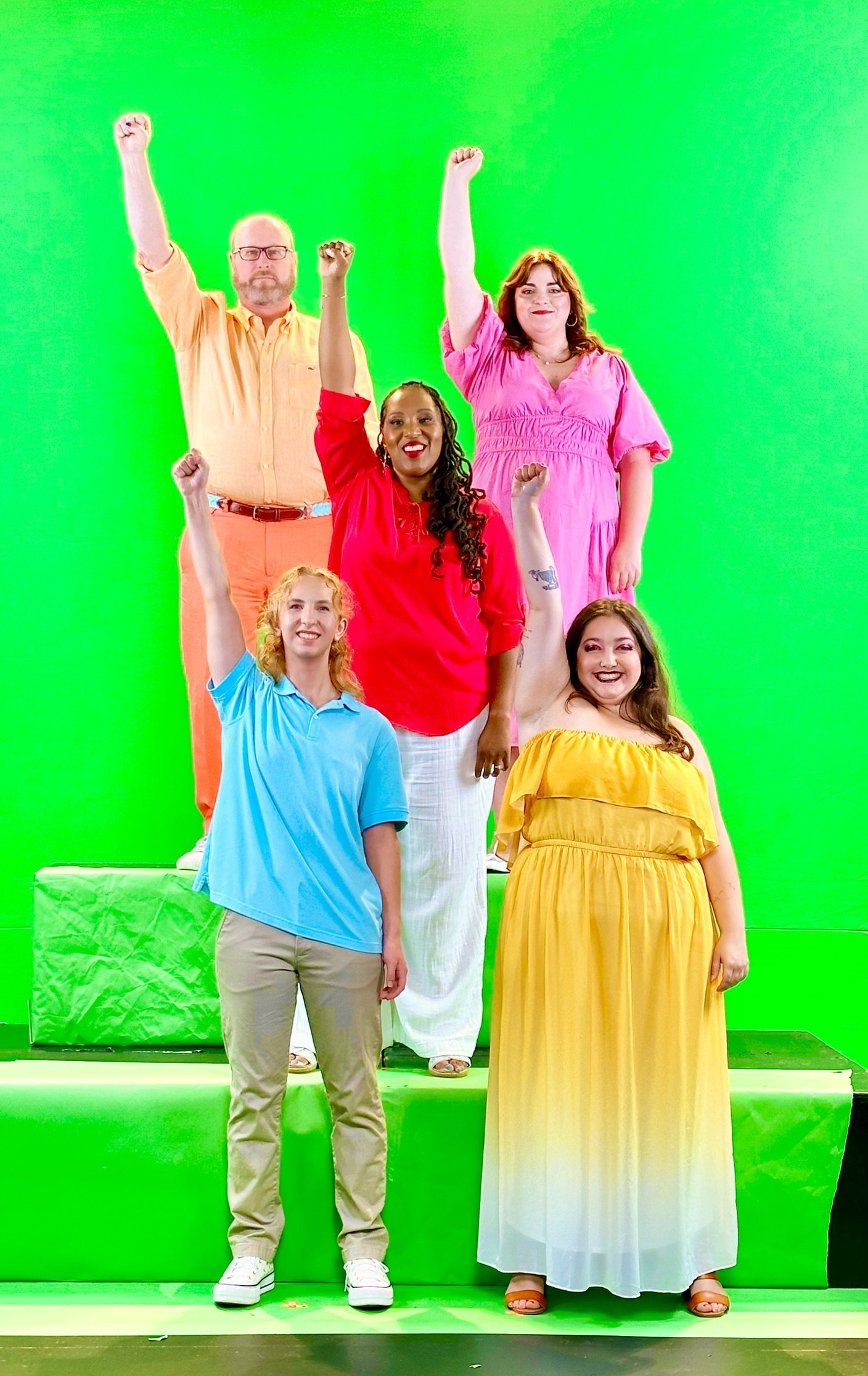 Six diverse people standing in front of a green screen, lifting their fists in the air, with one person in the front row wearing a yellow dress, the others in colorful casual outfits.