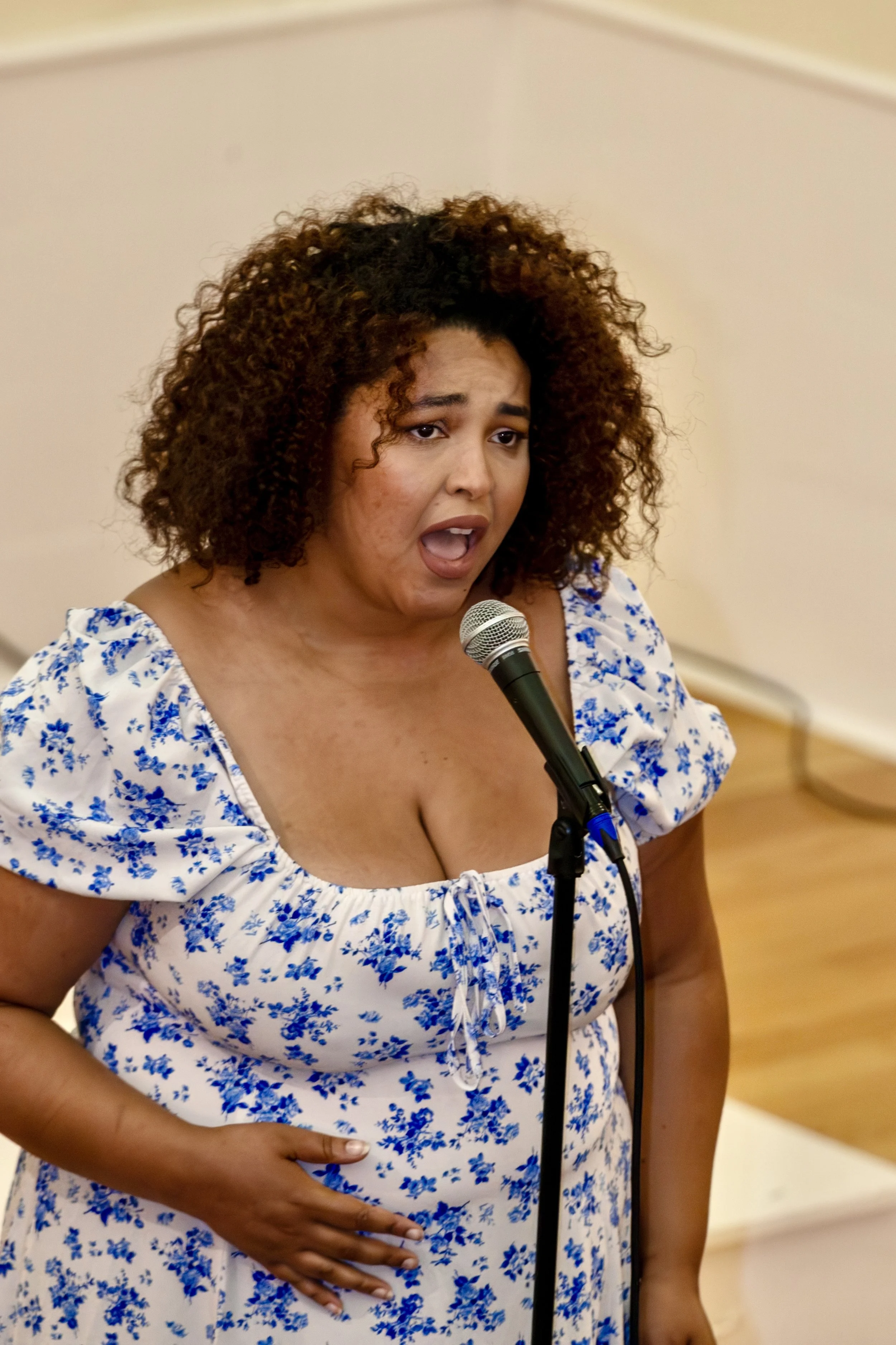 A woman with curly hair in a floral dress singing into a microphone.