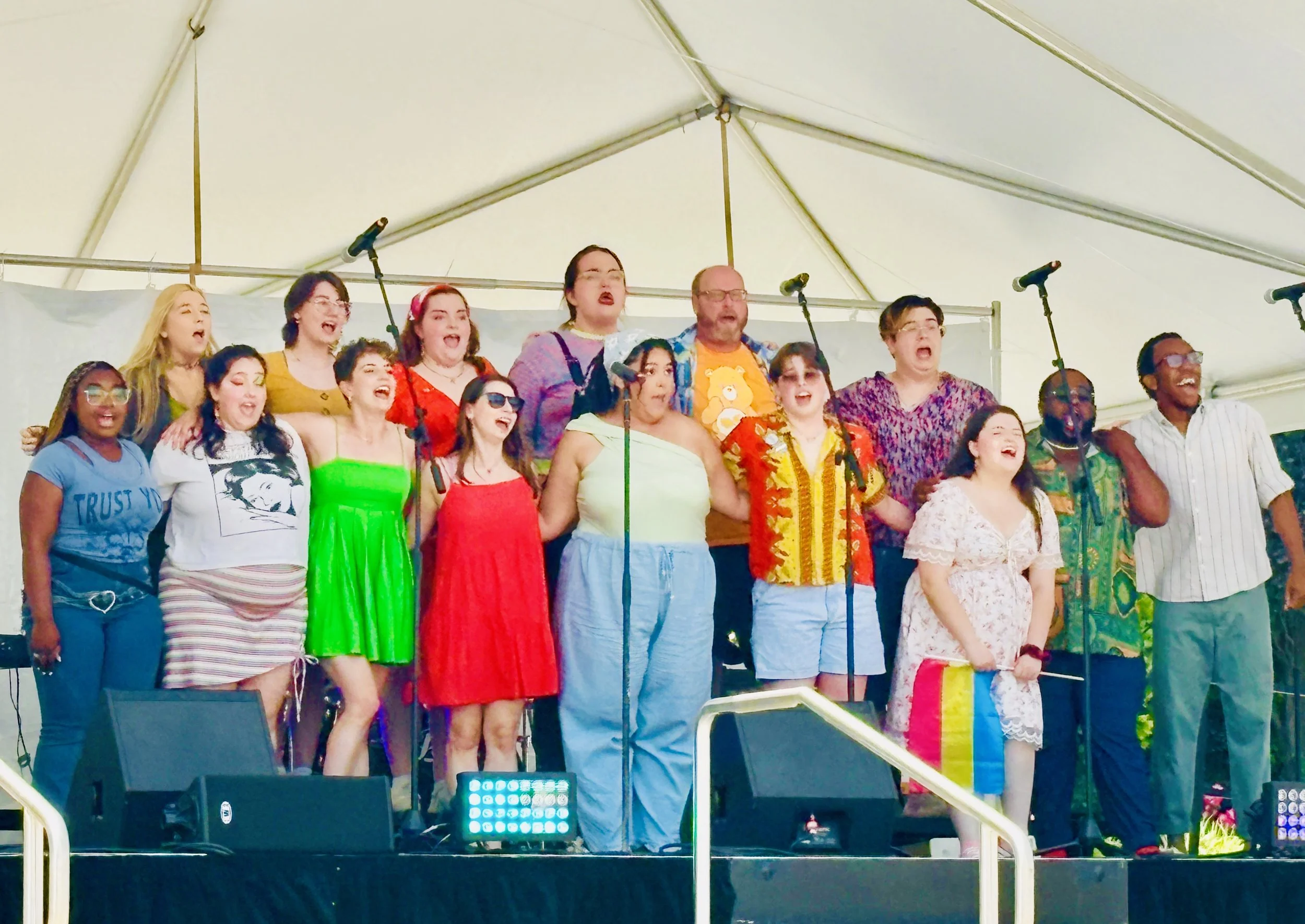 A diverse group of people singing together on a stage under a white canopy at an outdoor event.