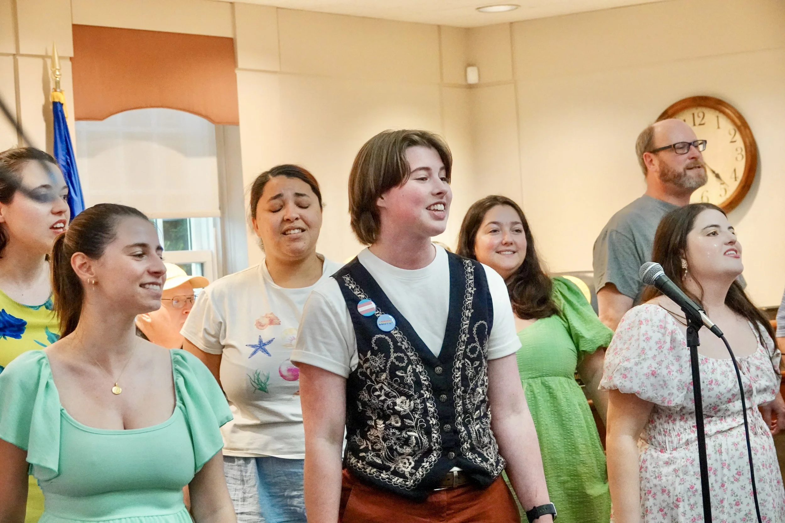 A group of people singing together in a room, standing in front of a microphone with a clock on the wall.