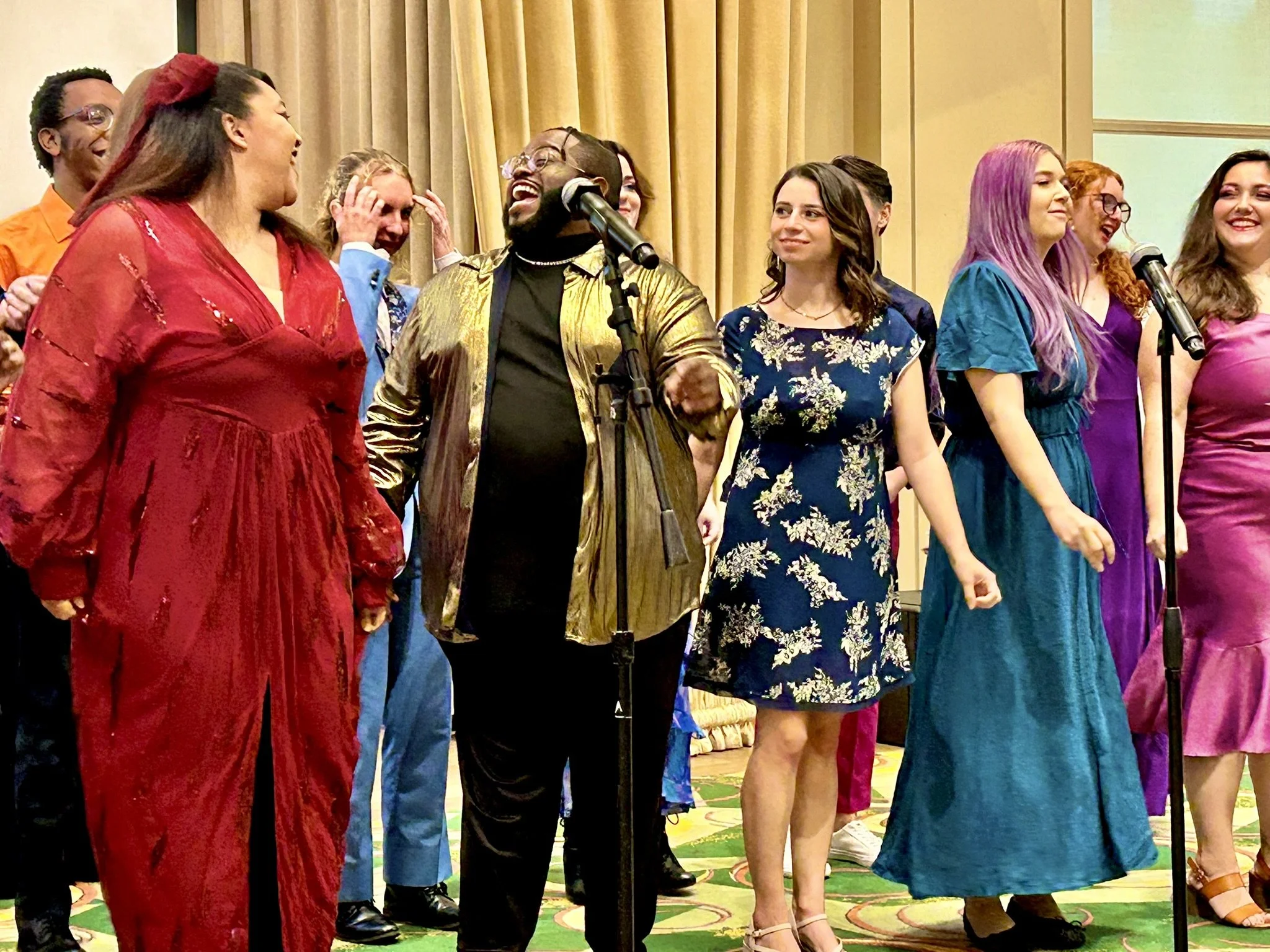 Group of diverse people laughing and smiling on stage, dressed in colorful dresses and suits, with some holding microphones.