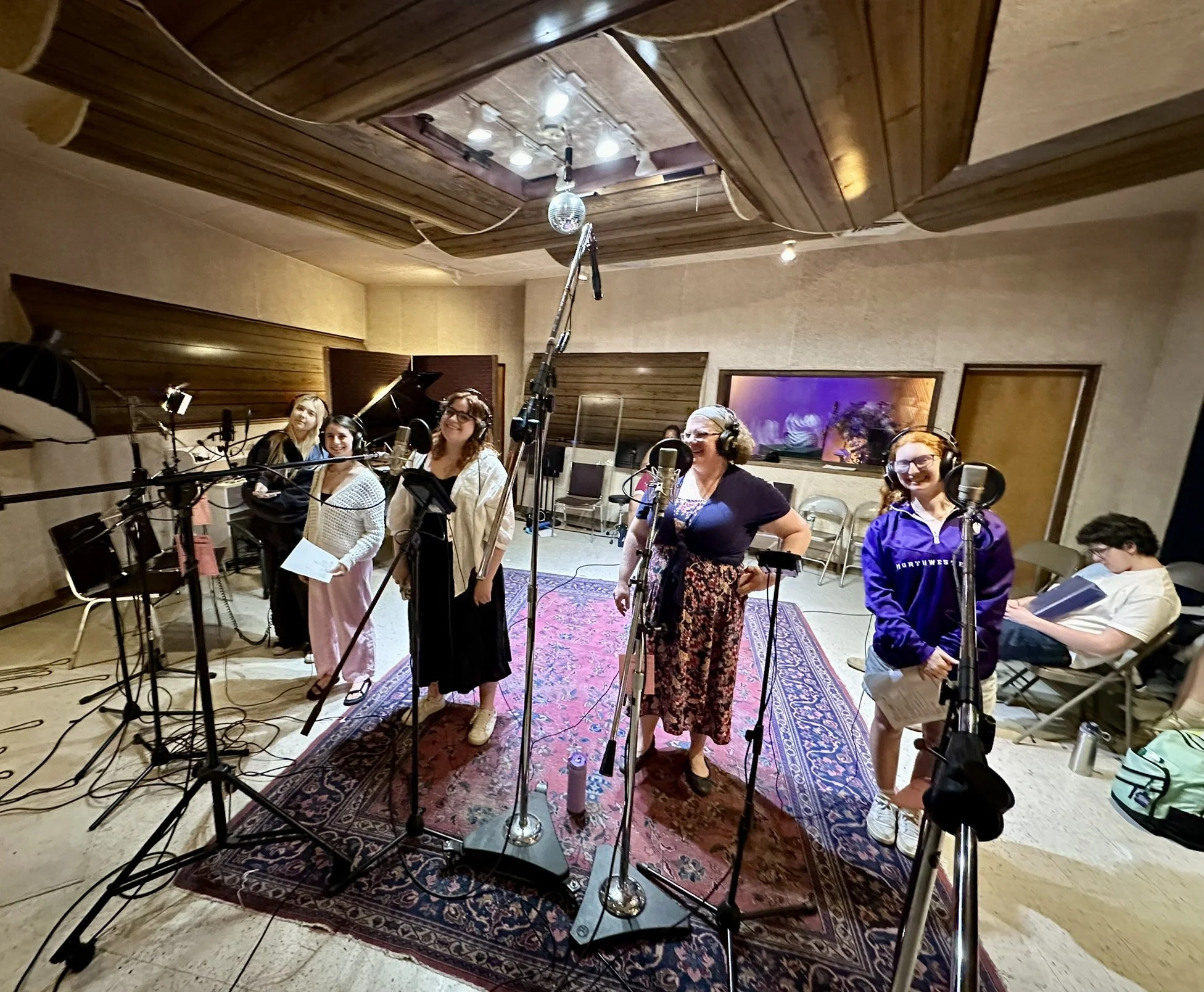 Group of women and a young girl in a recording studio with microphones, headphones, and music equipment, some standing and one sitting reading a script.