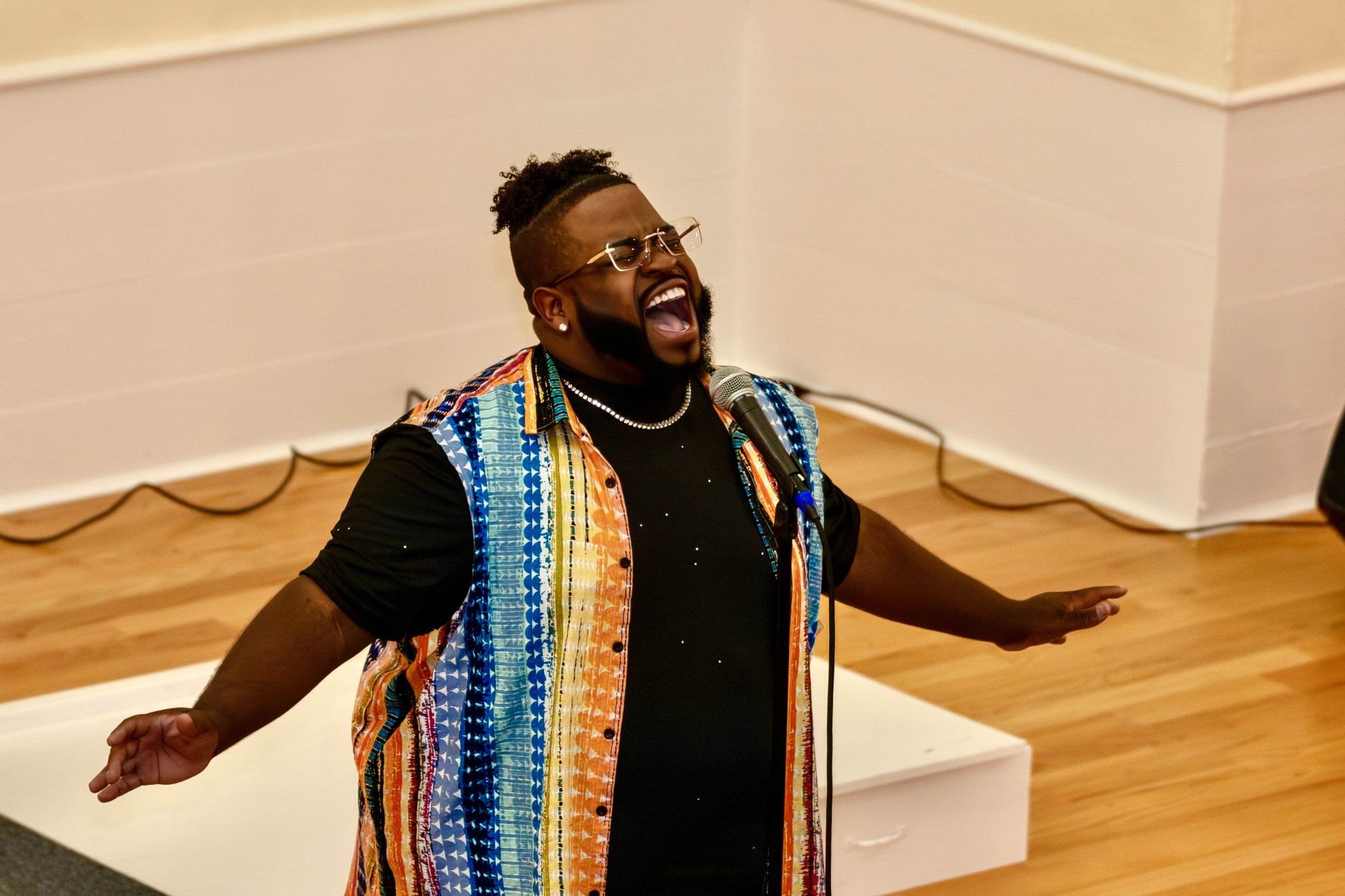 A man singing passionately into a microphone with his arms outstretched in a room with wooden flooring and white walls.