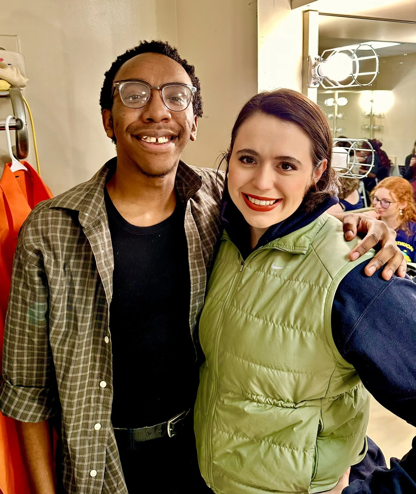 Two smiling people posing together, man with glasses and a woman in a green vest, in an indoor setting.