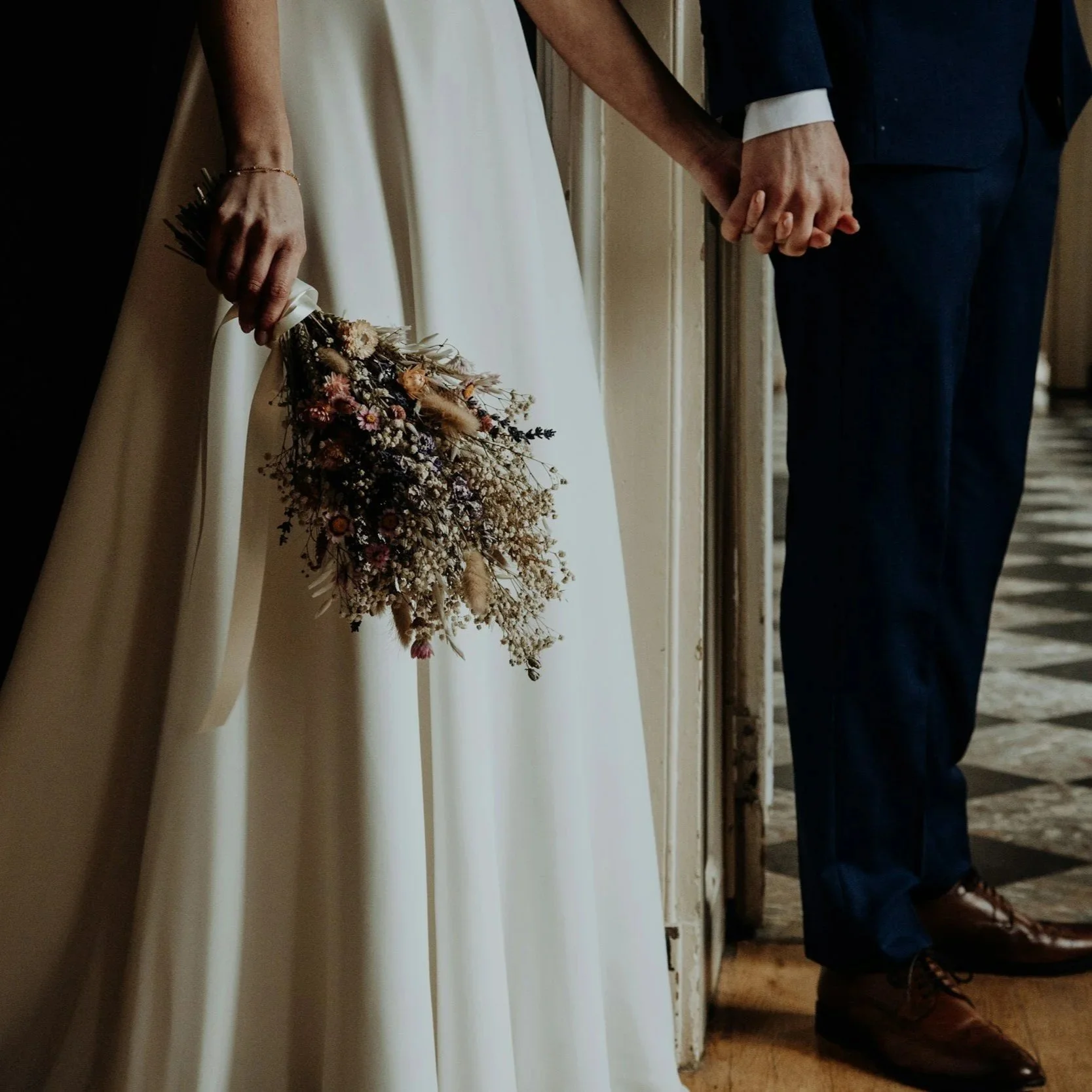 The image shows a close-up of a bride and groom holding hands, with the bride holding a bouquet of dried flowers. The bride is wearing a white dress, and the groom is dressed in a dark suit with a white shirt. The scene appears to be at a wedding.