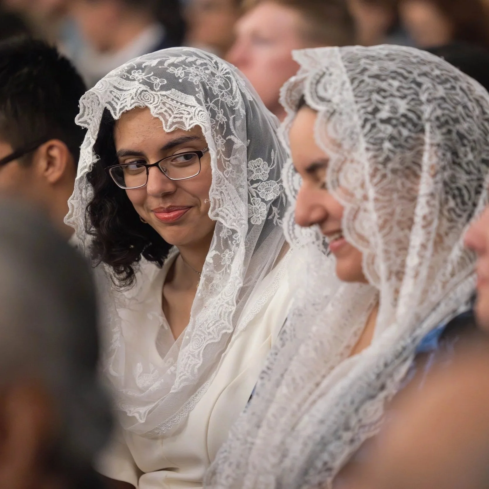Two women wearing white lace veils sit next to each other, one smiling and one with a neutral expression in a crowded indoor setting.