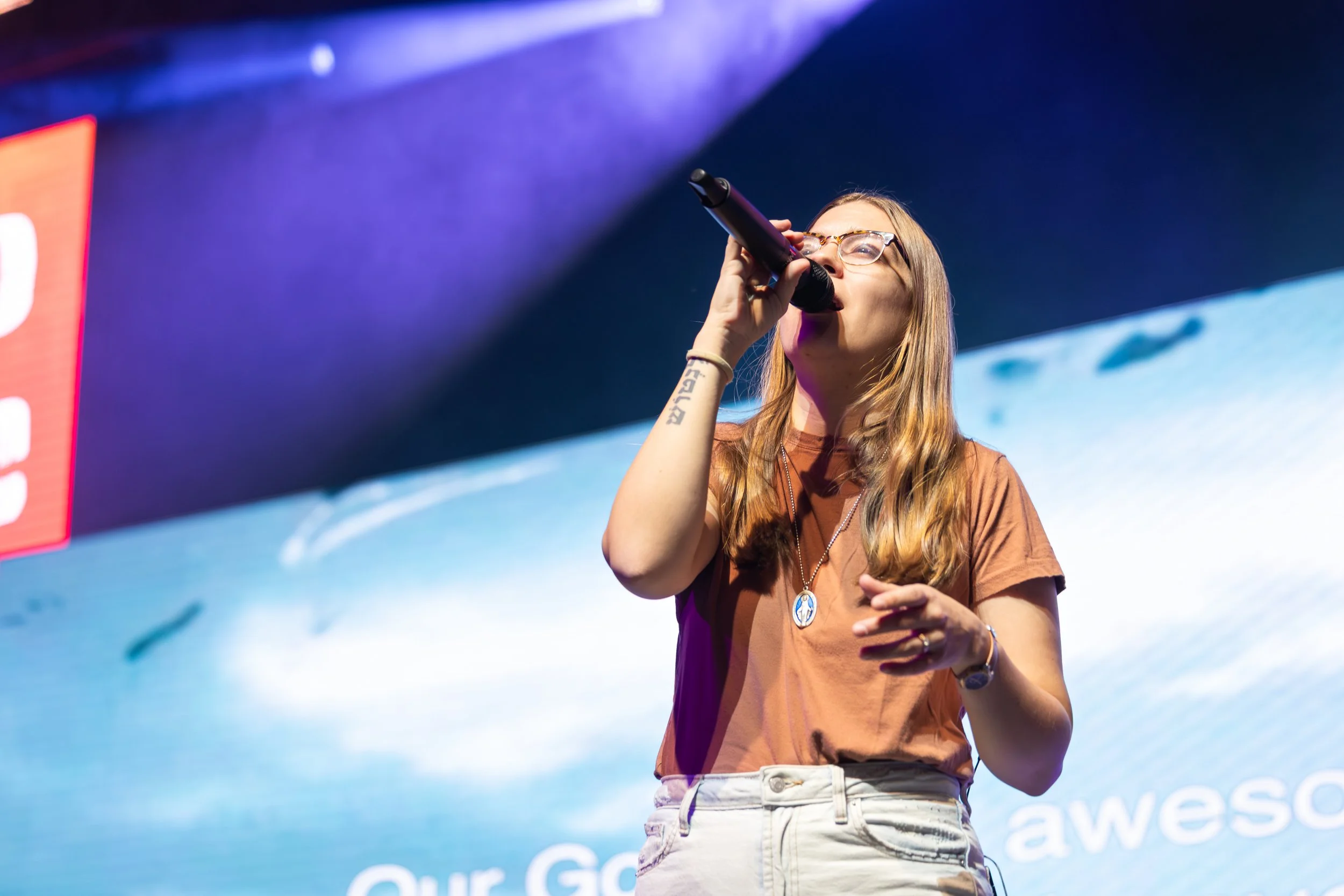 A young woman with glasses and long hair singing into a microphone on stage with LED screen in the background.