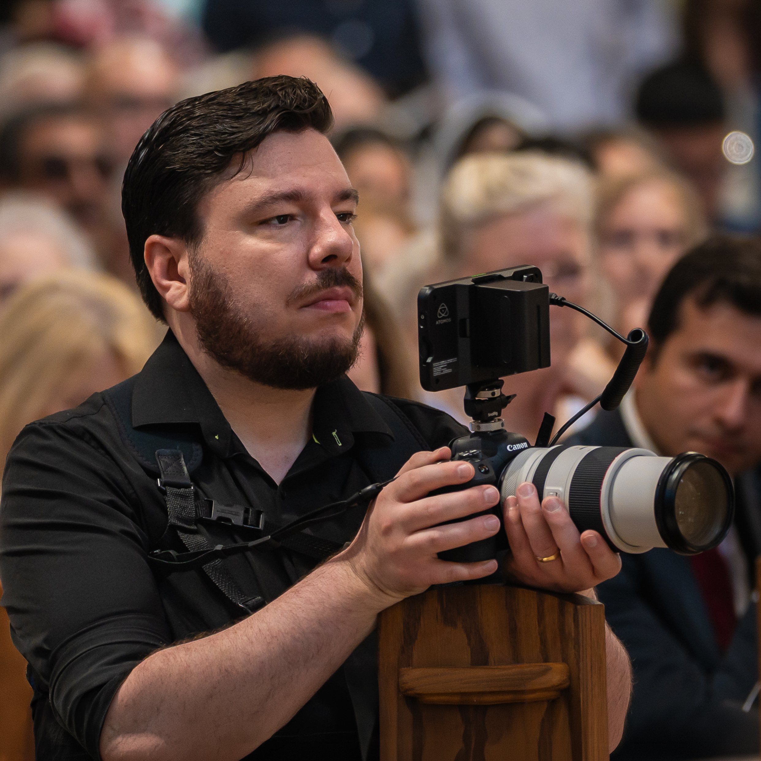 A man with dark hair and a beard using a professional camera in a crowded indoor setting.