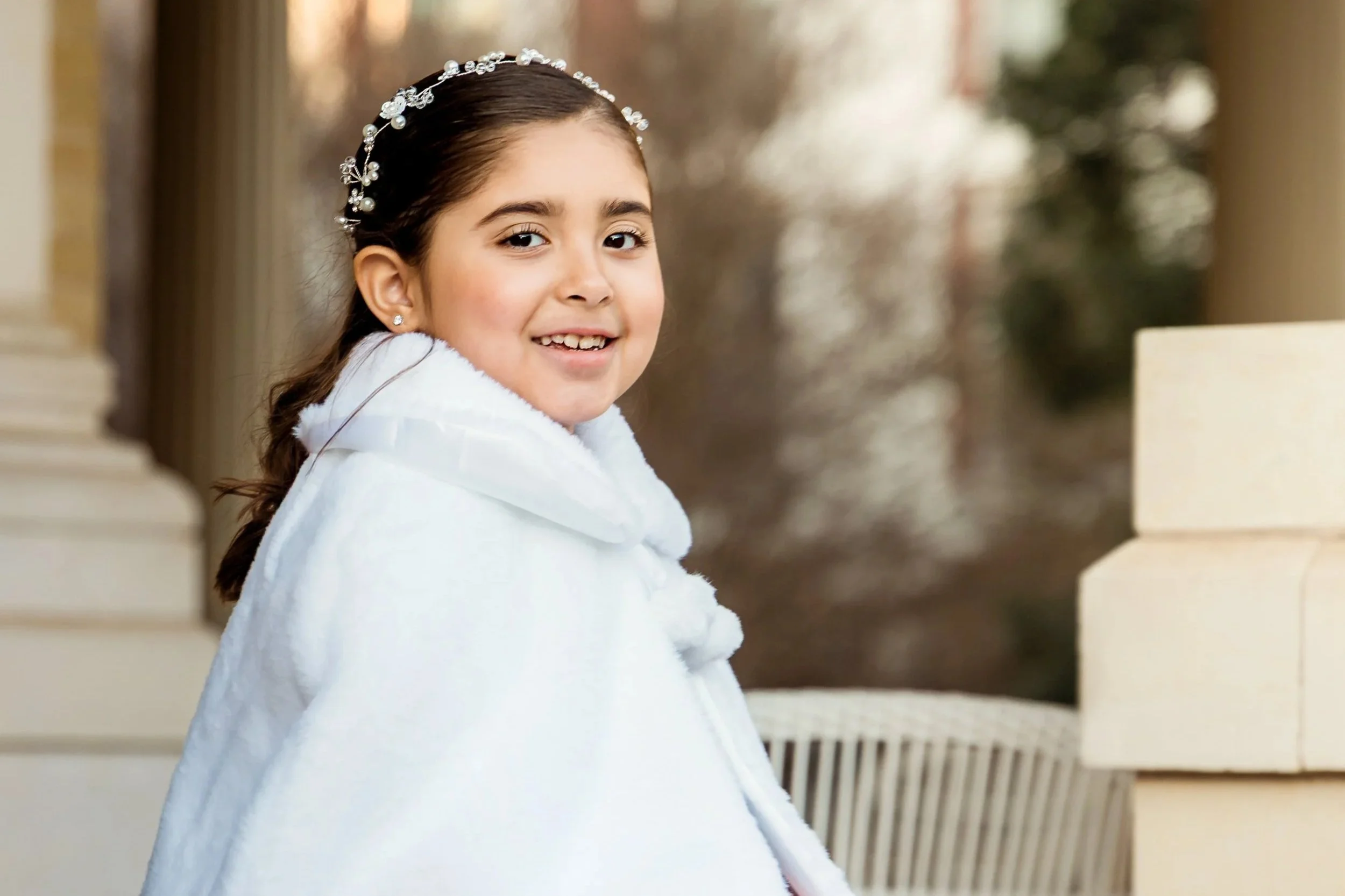 A young girl with dark hair and light skin smiling, wearing pearl jewelry and a white coat, standing on a balcony.
