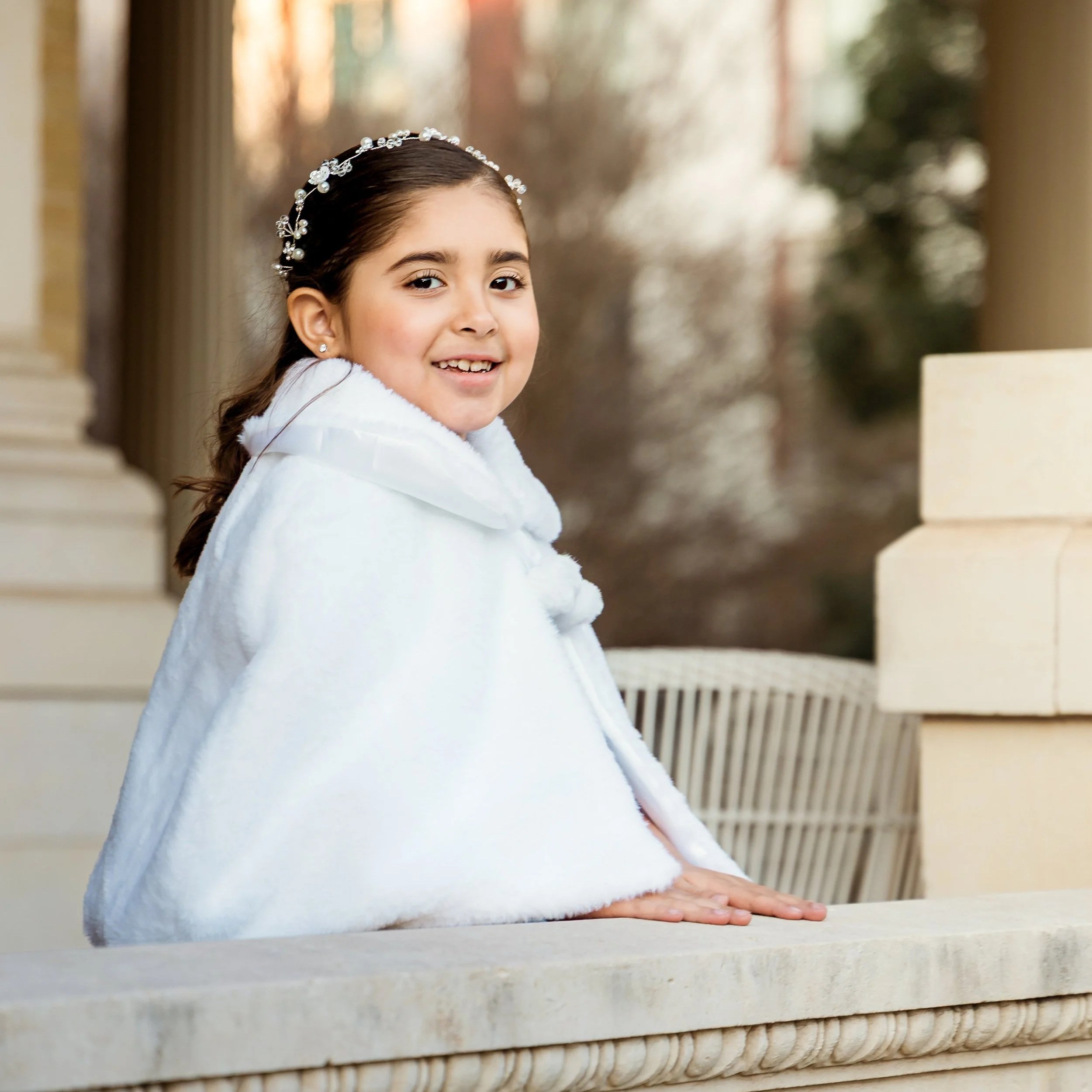 A young girl with dark hair styled in a braid, wearing a white fuzzy coat and a decorative headband with pearls and flowers, smiling as she leans on a white surface.