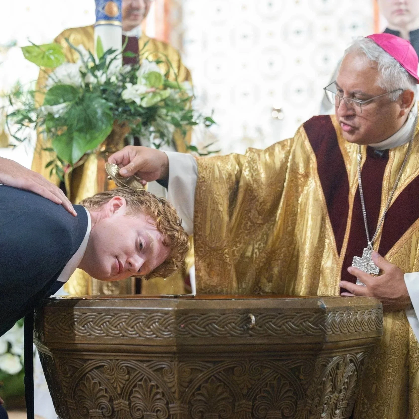 A religious ceremony where a bishop in gold robes anoints a man with oil on his forehead, with a priest in matching robes overseeing the event, in front of a decorated background with flowers.