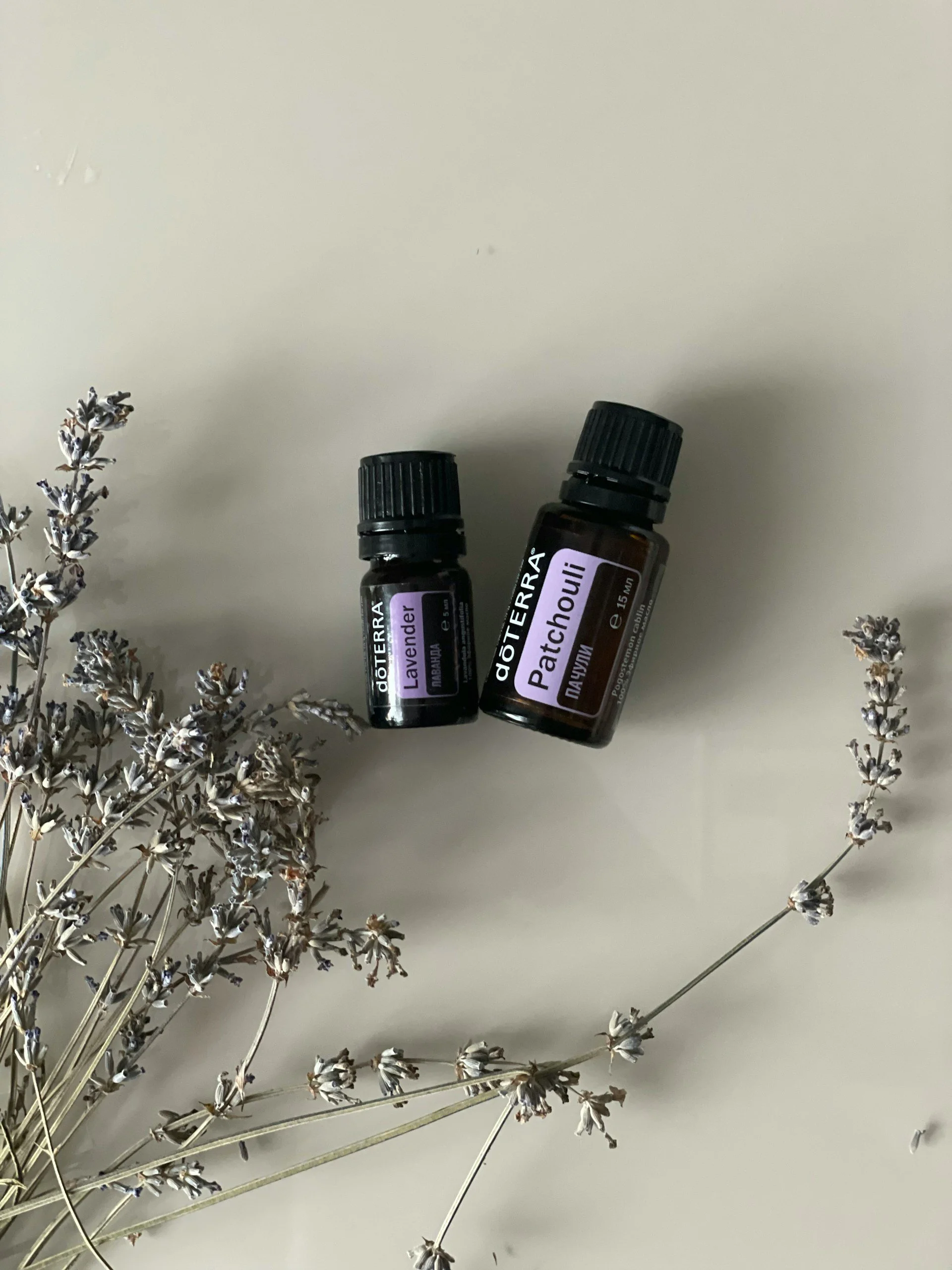 Twee flesjes etherische olie van doTERRA, lavendel en patchouli, liggen naast gedroogde lavendel op een lichtkleurige tafel.