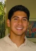 A young man with dark hair smiling, wearing a light-colored shirt, standing indoors with a green poster and plant in the background.