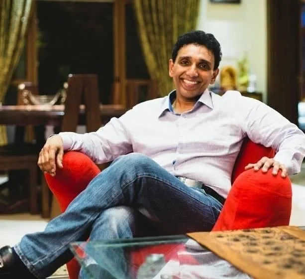 A man sitting on a red armchair in a cozy room, smiling at the camera with crossed legs. The background features windows with curtains and a dining table.