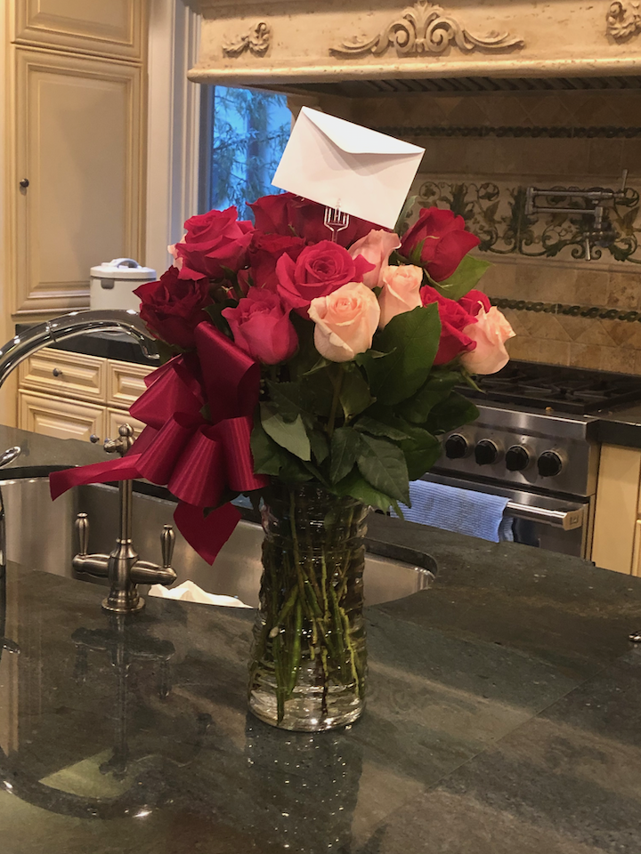 A glass vase with a bouquet of red, pink, and peach roses, decorated with a red bow and a white envelope, placed on a kitchen counter.