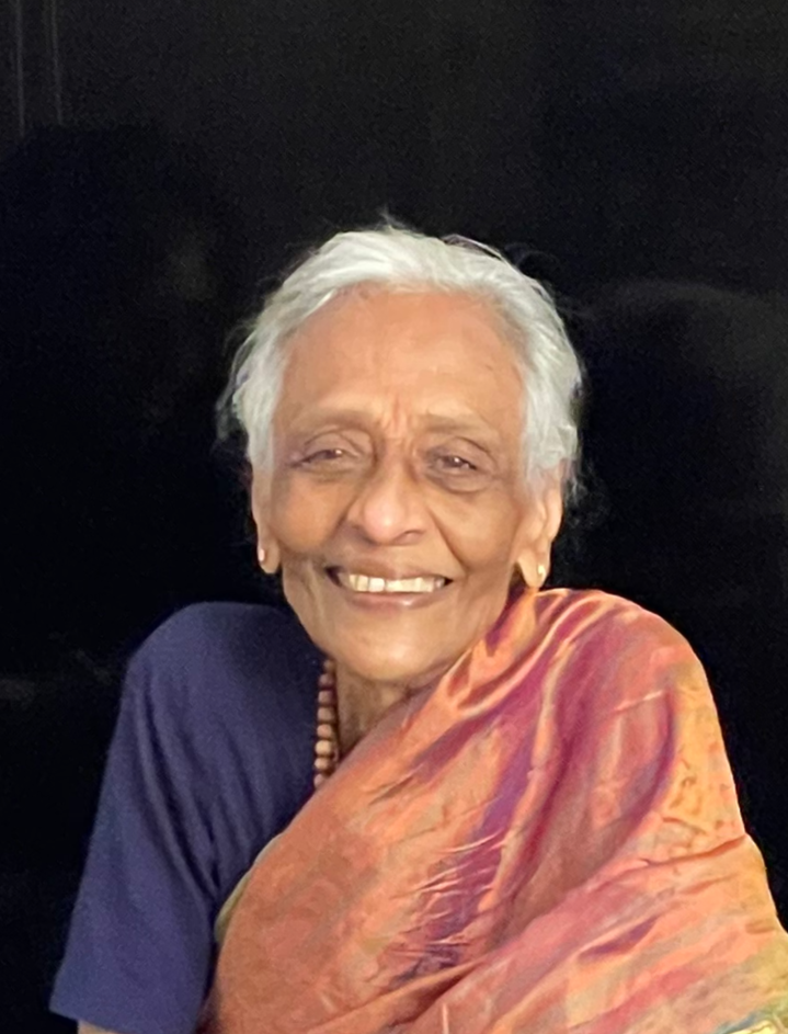 An elderly woman with white hair smiling at the camera, wearing a blue top and a colorful shawl.