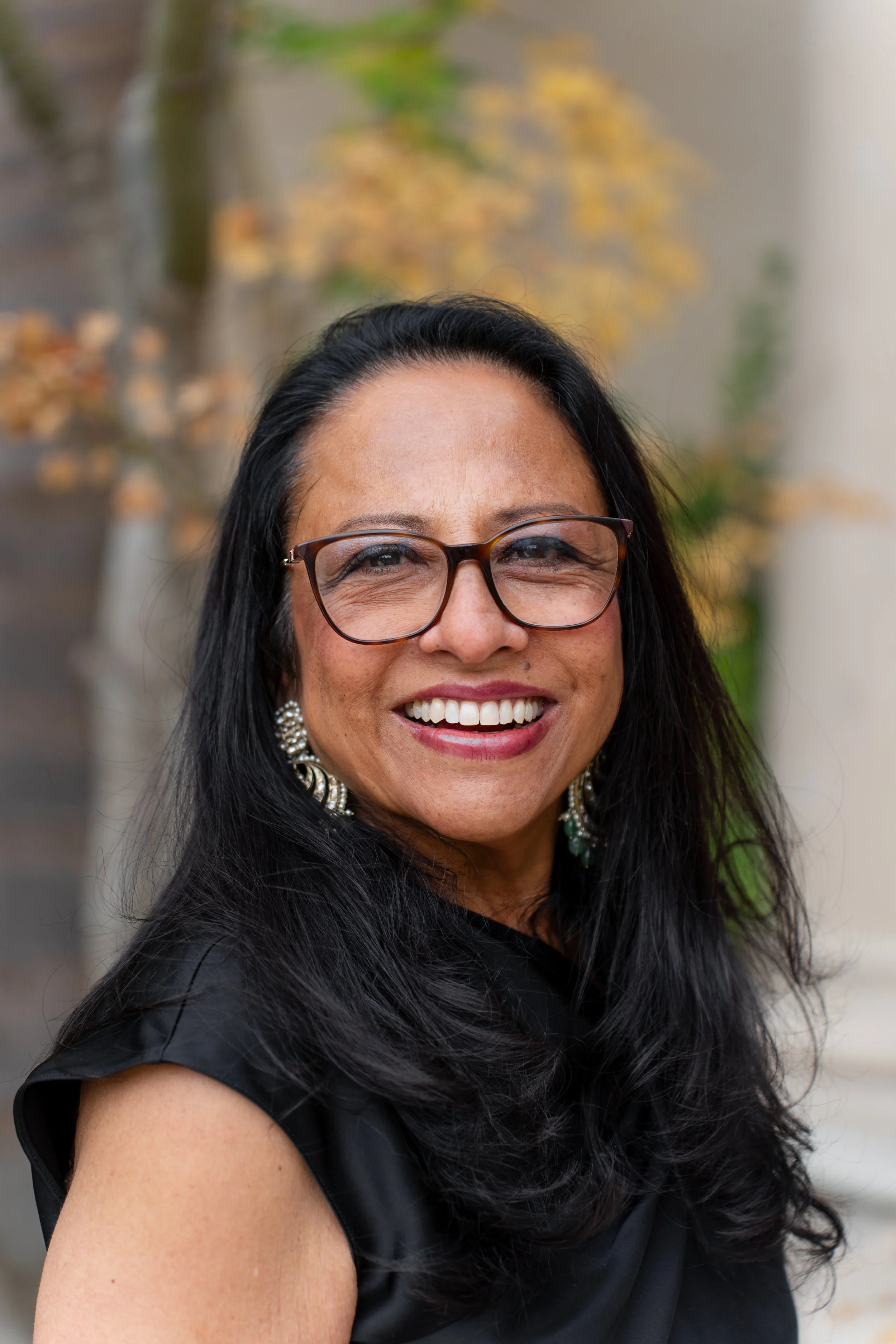 A woman with long black hair, glasses, and earrings, smiling outdoors with blurred foliage in the background.