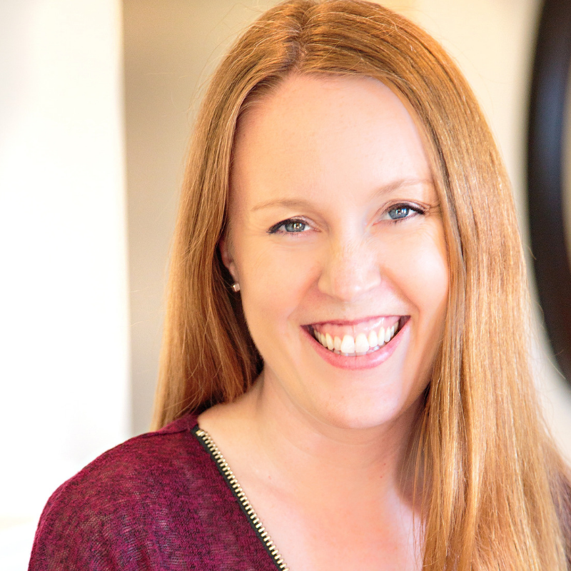 Smiling woman with long red hair wearing a burgundy top near a mirror