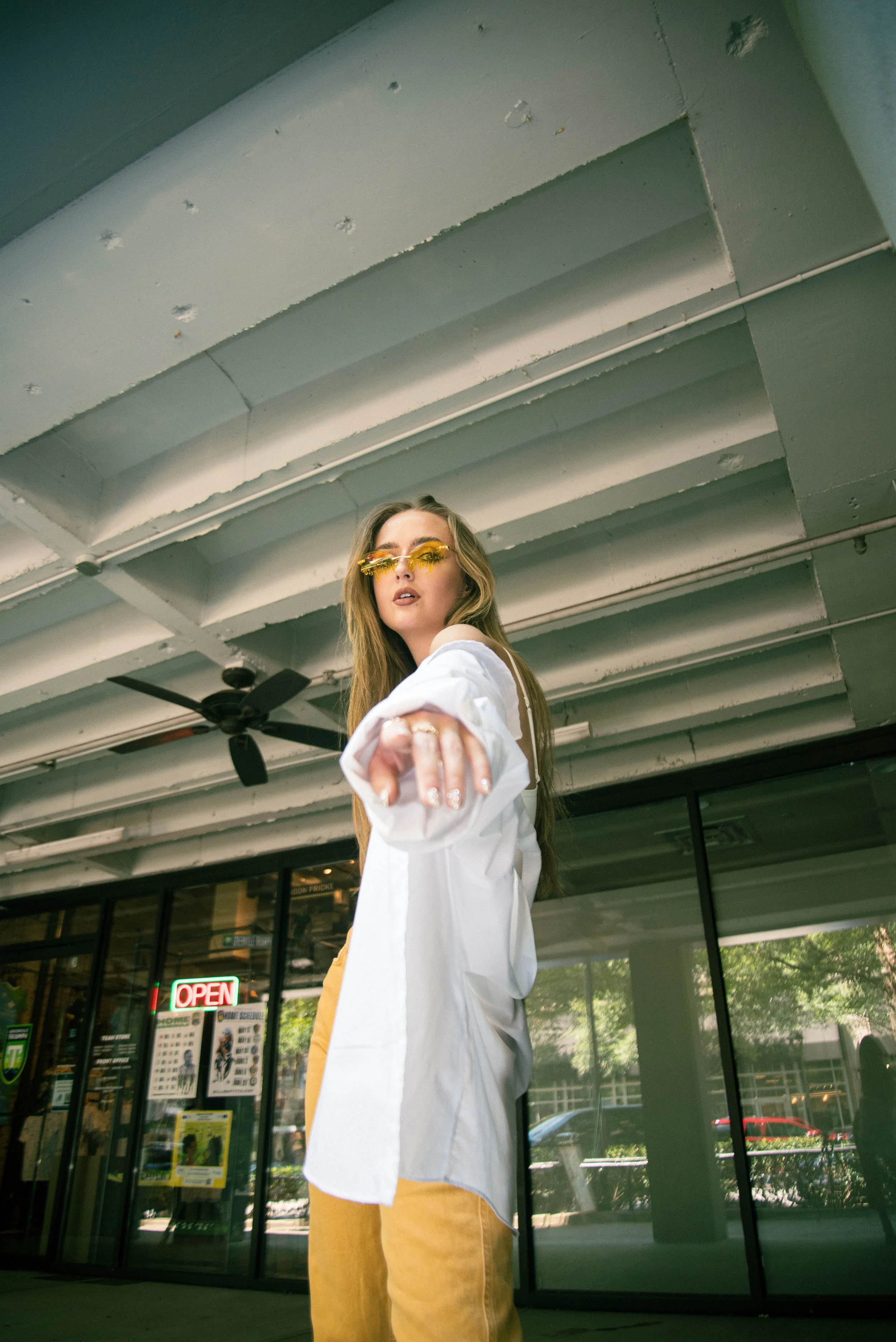 A young woman with long hair wearing yellow-tinted sunglasses, a white long-sleeve shirt, and mustard-colored pants standing in front of a store entrance with an "Open" sign in the window, a ceiling with exposed beams, and a ceiling fan.