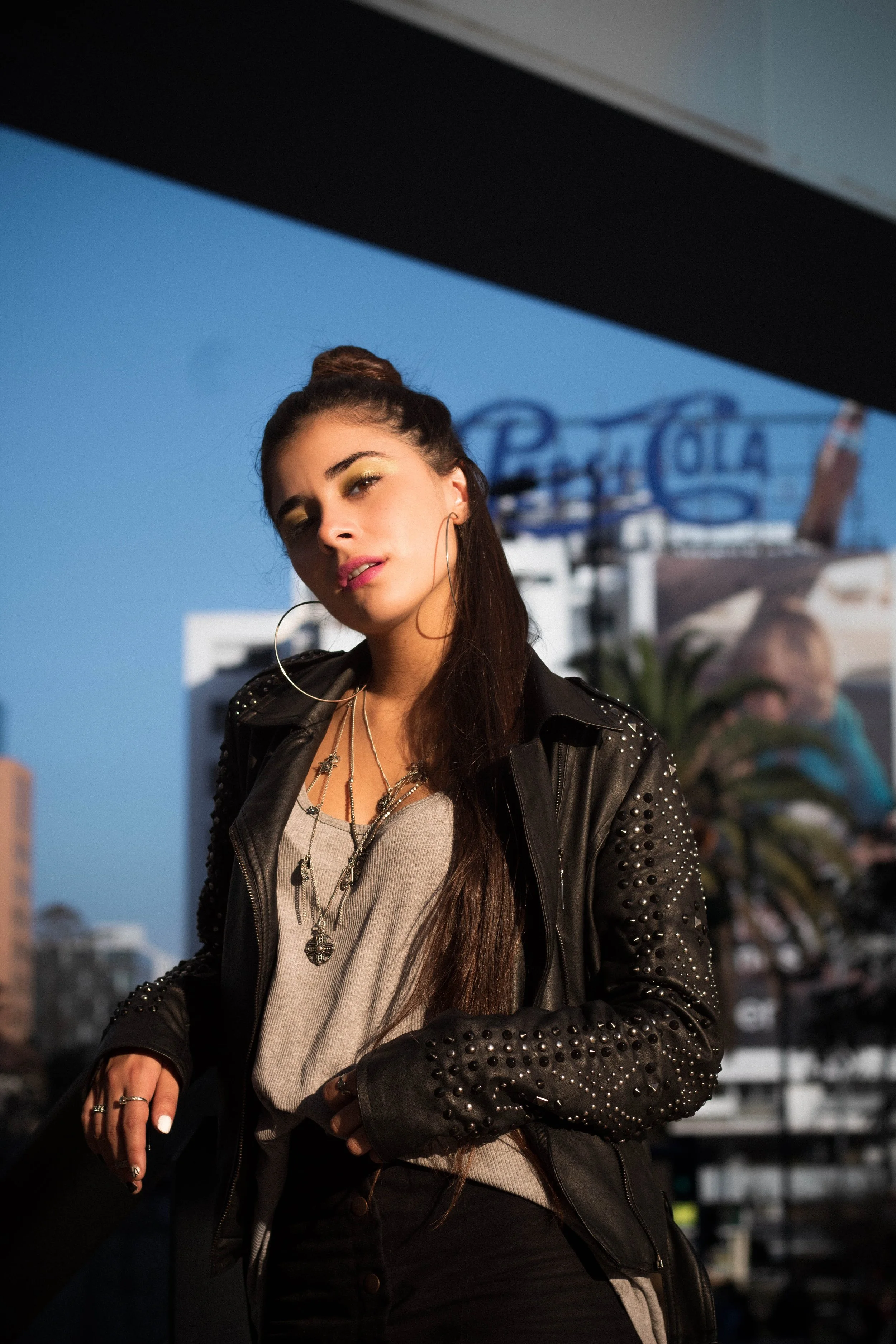 A young woman with long dark hair styled in a top knot, wearing a leather jacket with studded detail, a beige top, multiple necklaces, and large hoop earrings, standing outdoors with a blue sky and a blurred sign in the background.