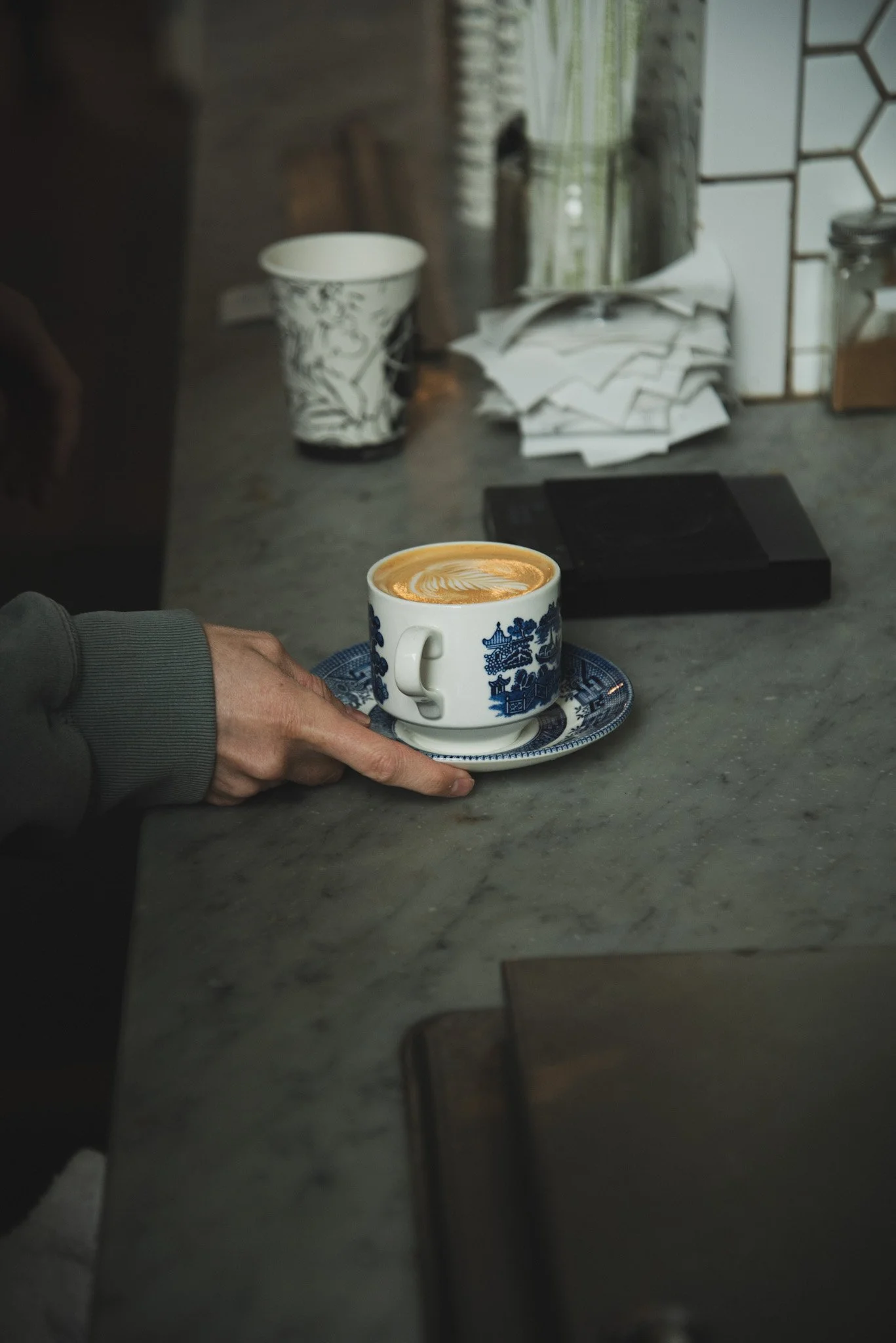 A person's hand holding a small blue and white ceramic cup with a latte art heart design, resting on a matching saucer on a gray marble countertop. In the background, there are disorganized papers, a black electronic device, a white patterned cup, a 