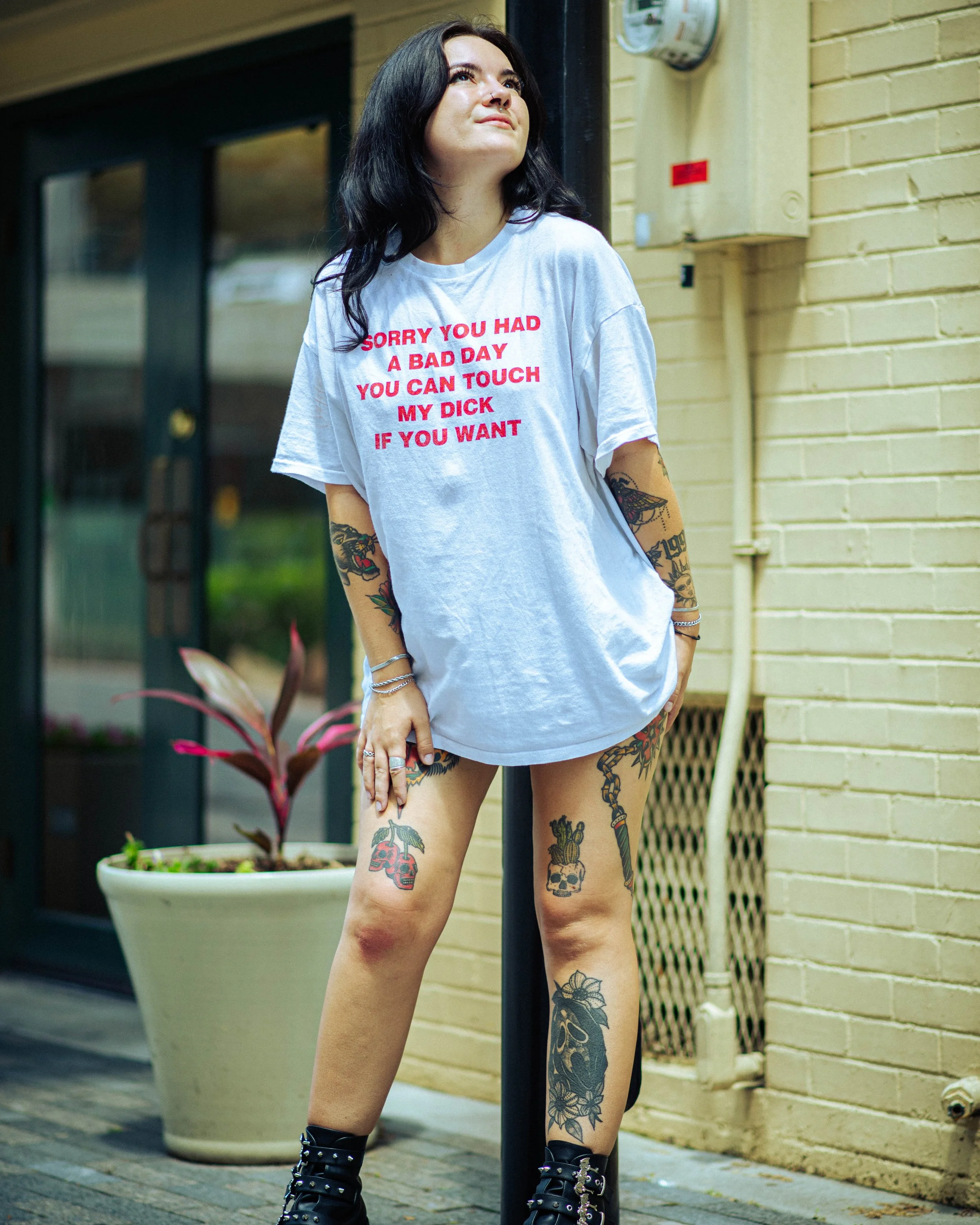 A woman with black hair, tattoos on her arms and legs, wearing a white t-shirt with red text, black boots, standing outdoors next to a yellow brick wall and a street pole. The T-shirt has explicit language.