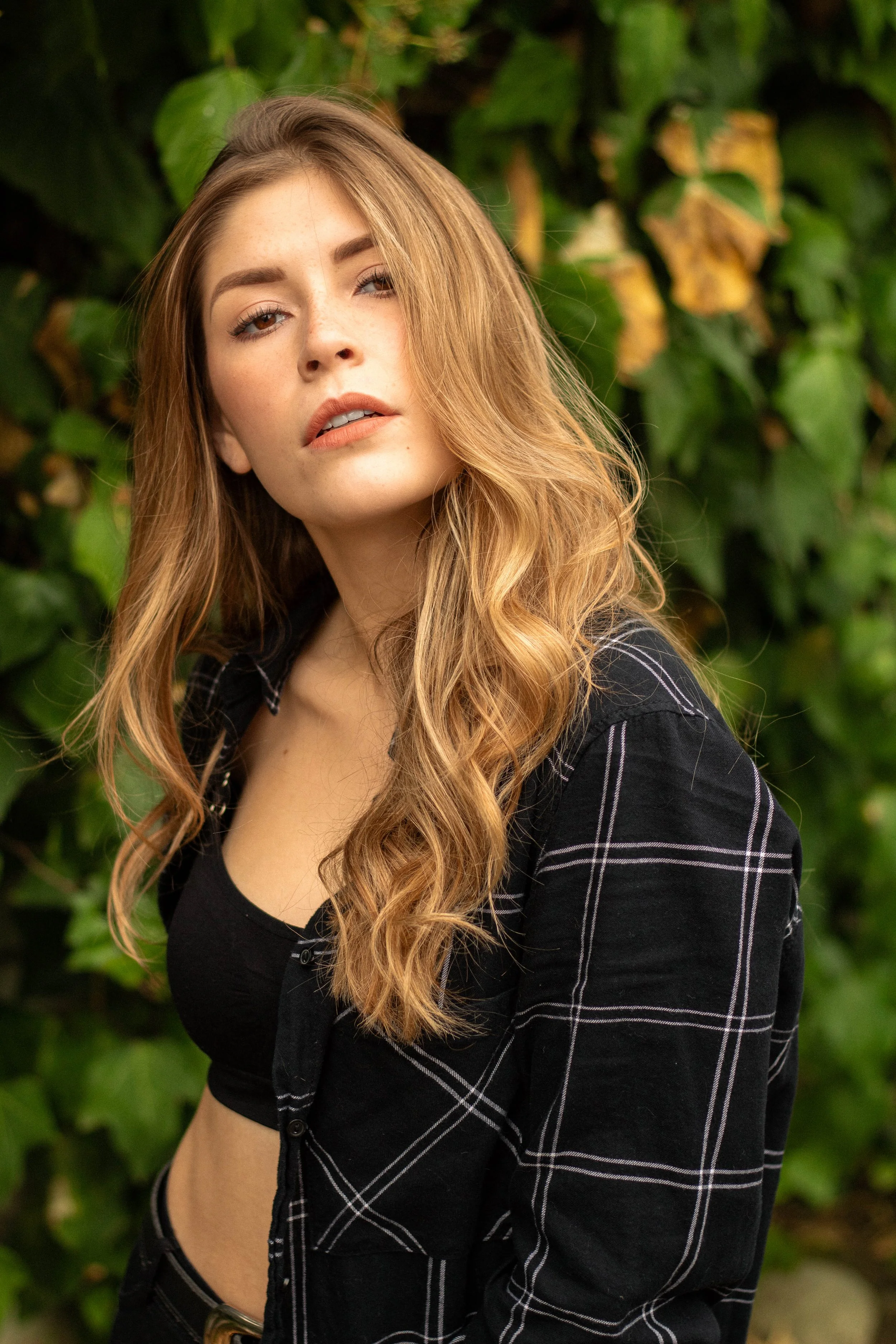A young woman with long, wavy red hair and light makeup, wearing a black shirt and a black plaid jacket, standing outdoors in front of green leafy plants.