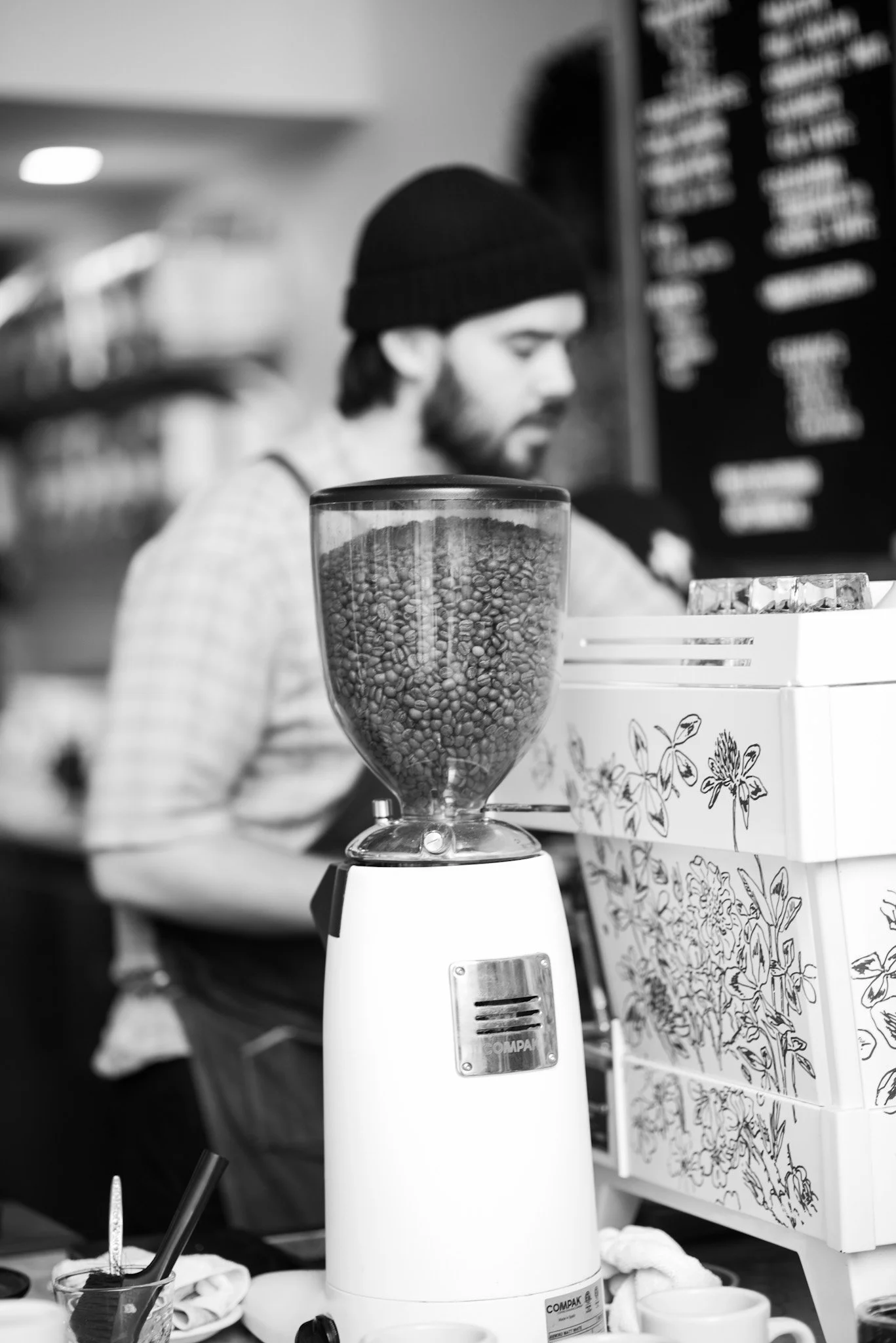 A person wearing a black beanie and a plaid shirt working behind a coffee grinder, with a blurred menu board in the background.