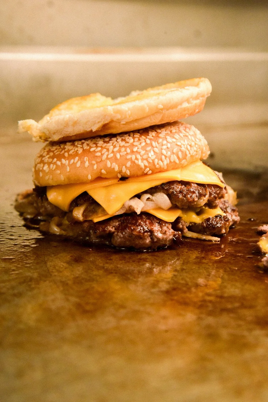 A partially assembled cheeseburger with a sesame seed bun, cheddar cheese, grilled beef patties, and onions, resting on a flat surface.