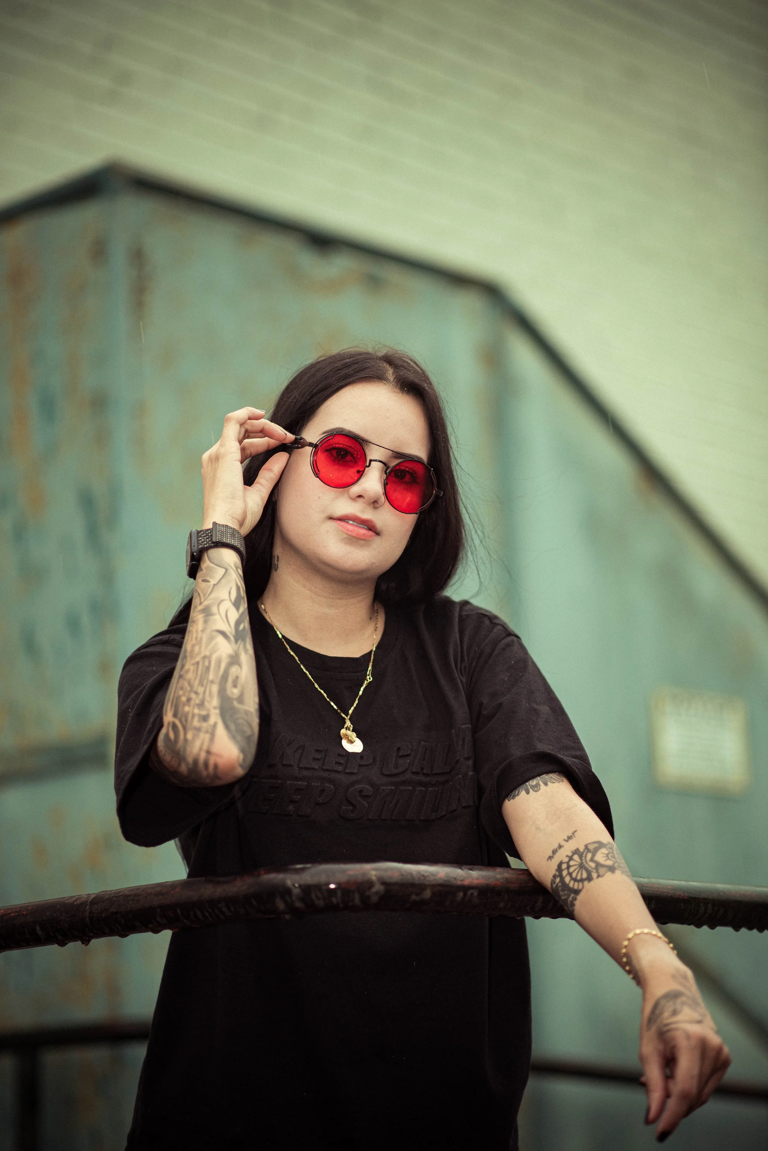 A woman with dark hair wearing red-tinted sunglasses, tattoos on her arms, a black t-shirt, and a gold necklace, sitting at a table with a rustic green wall in the background.