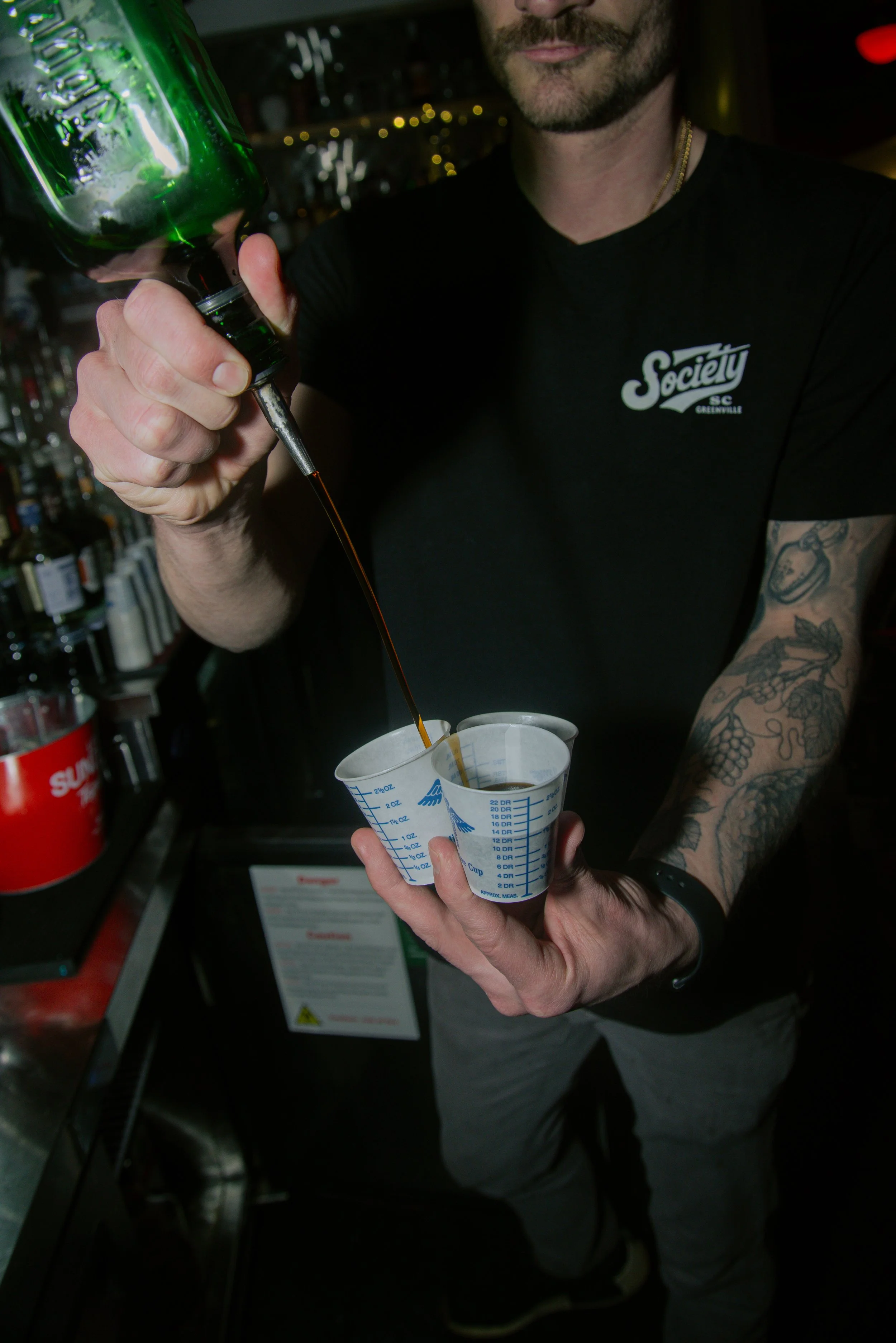 A man with a beard and tattoos on his arm is pouring a dark liquid into a paper measuring cup at a bar. He is wearing a black t-shirt with a logo that says 'Society SC Greenville' and a watch on his wrist. The background has shelves with bottles and 